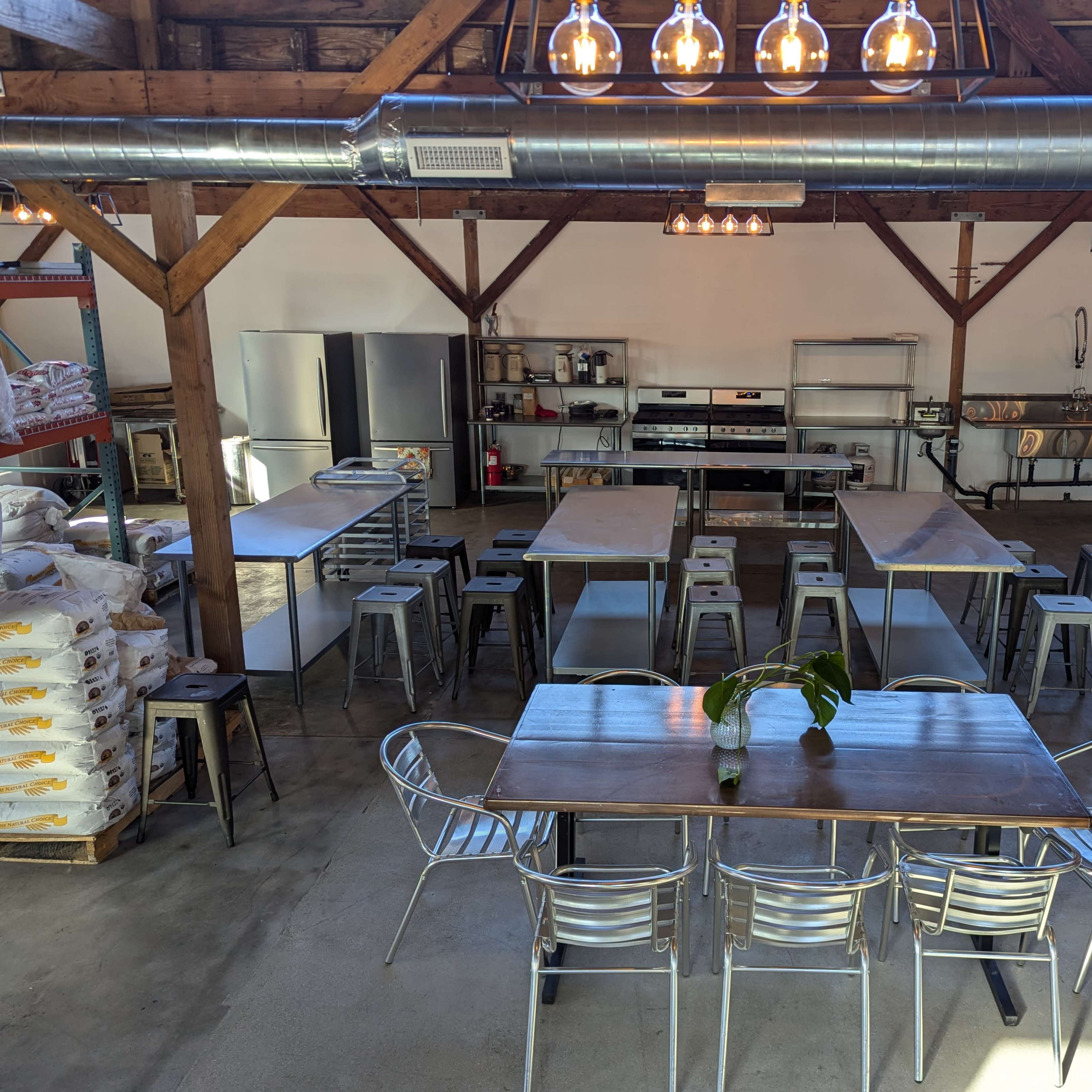 A spacious industrial kitchen setup with multiple stainless steel workstations, tables, and stools, illuminated by overhead lights.