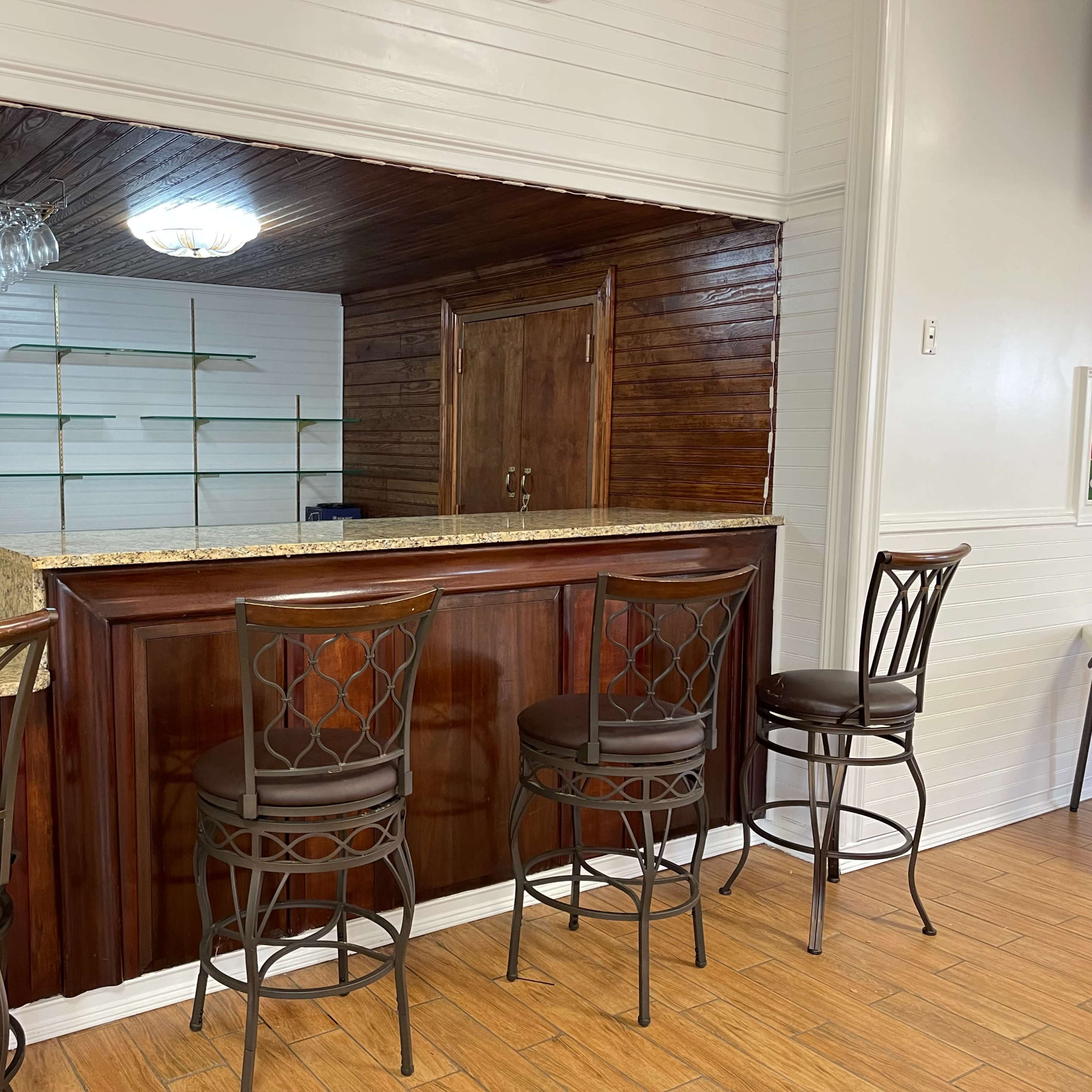 A small bar area with four high stools, a granite countertop, and a wooden back wall featuring shelves.