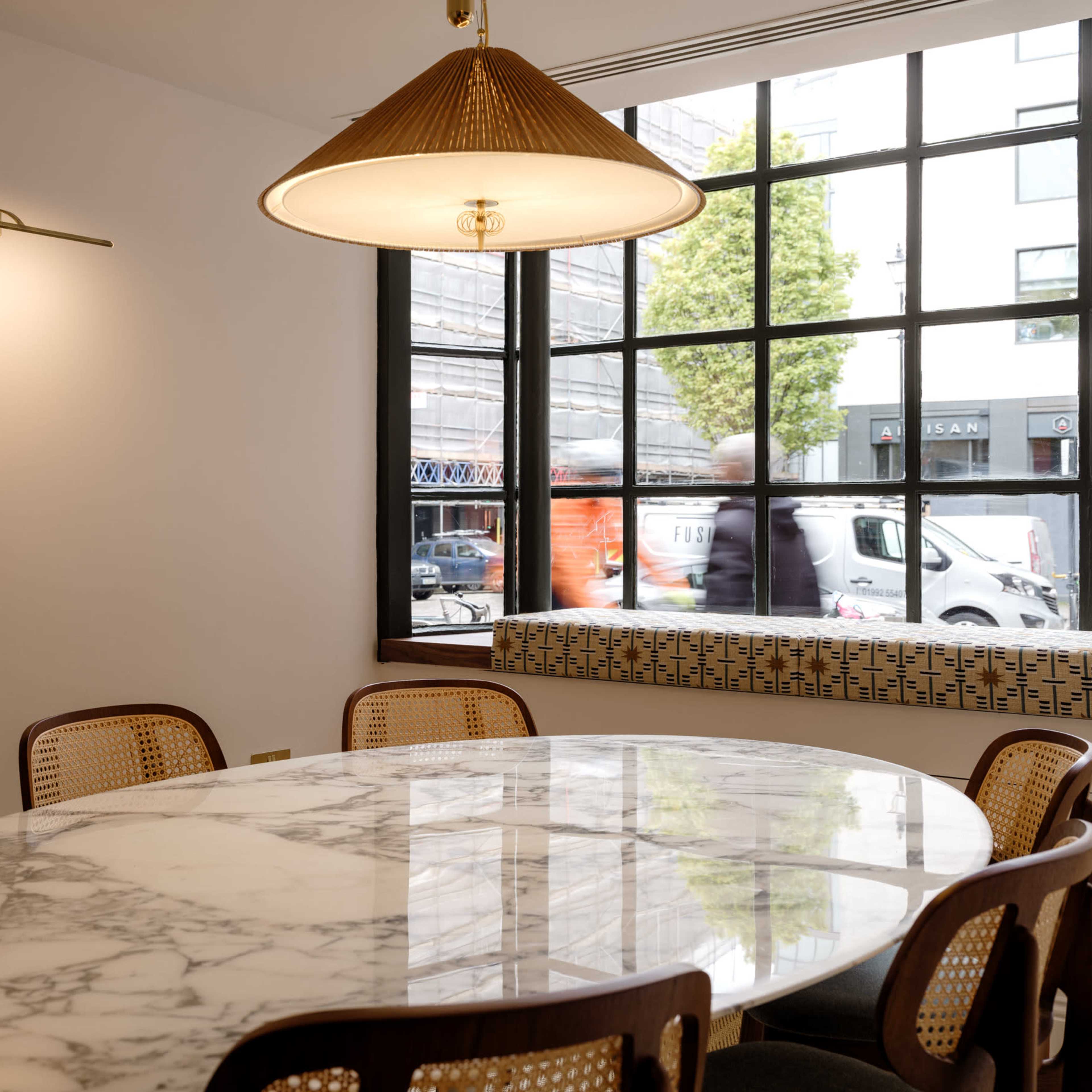 A round marble table surrounded by cane-back chairs is illuminated by a pendant light in a room with a large window displaying a street view.
