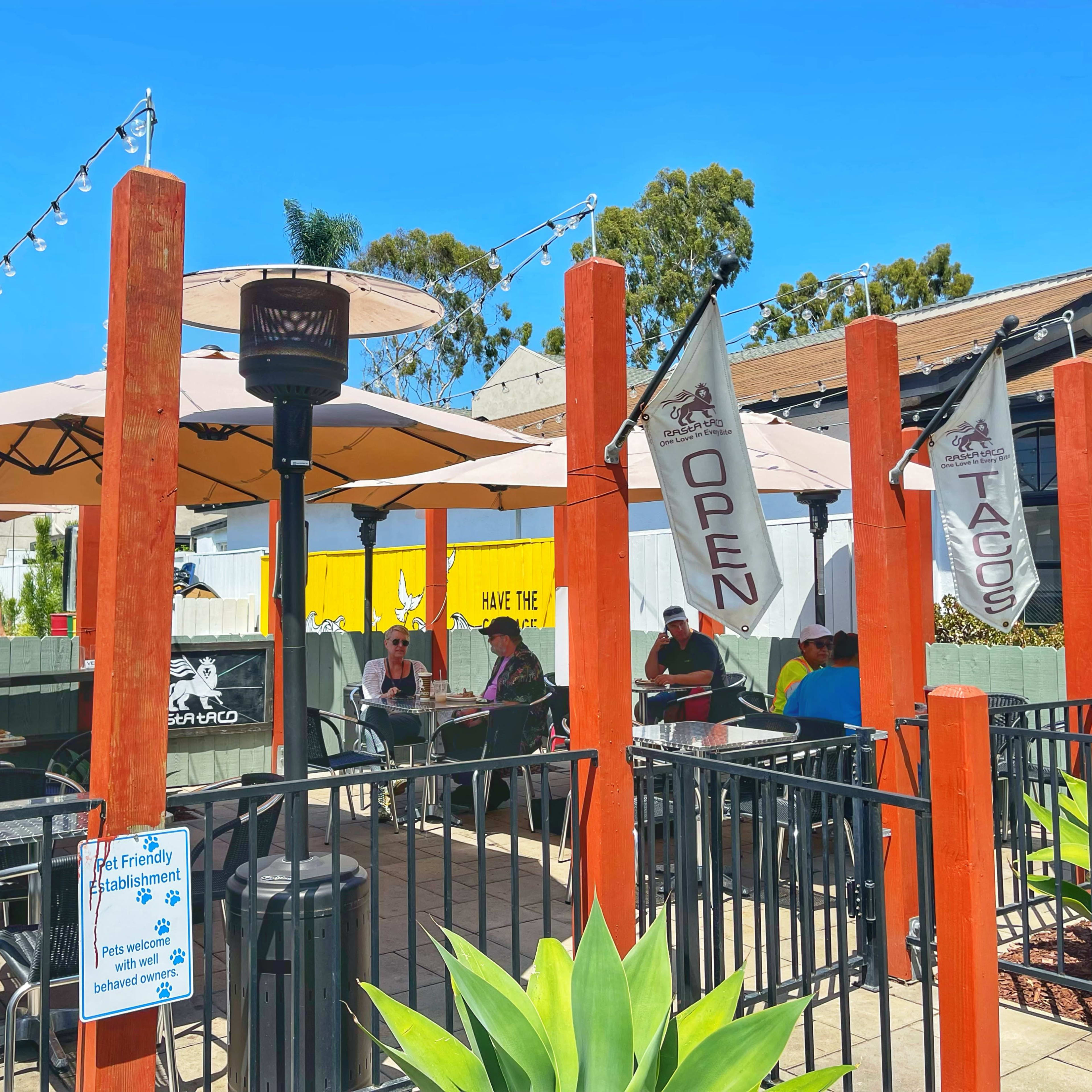 A bustling outdoor patio at a restaurant features diners seated under umbrellas, with flags displaying "Open" and "Tacos" prominently displayed.