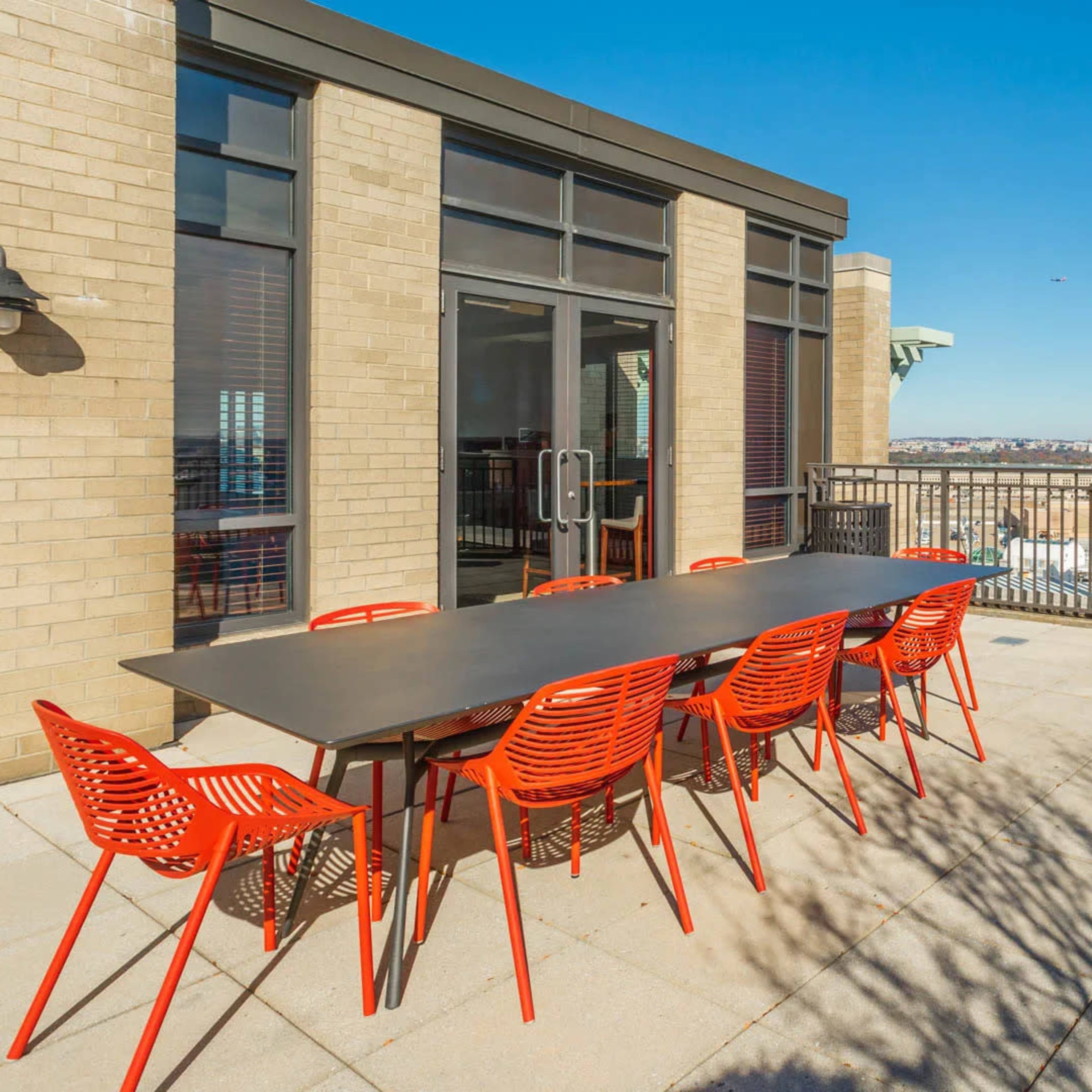 A rooftop patio features a large table with red chairs and a grill, overlooking the city skyline.