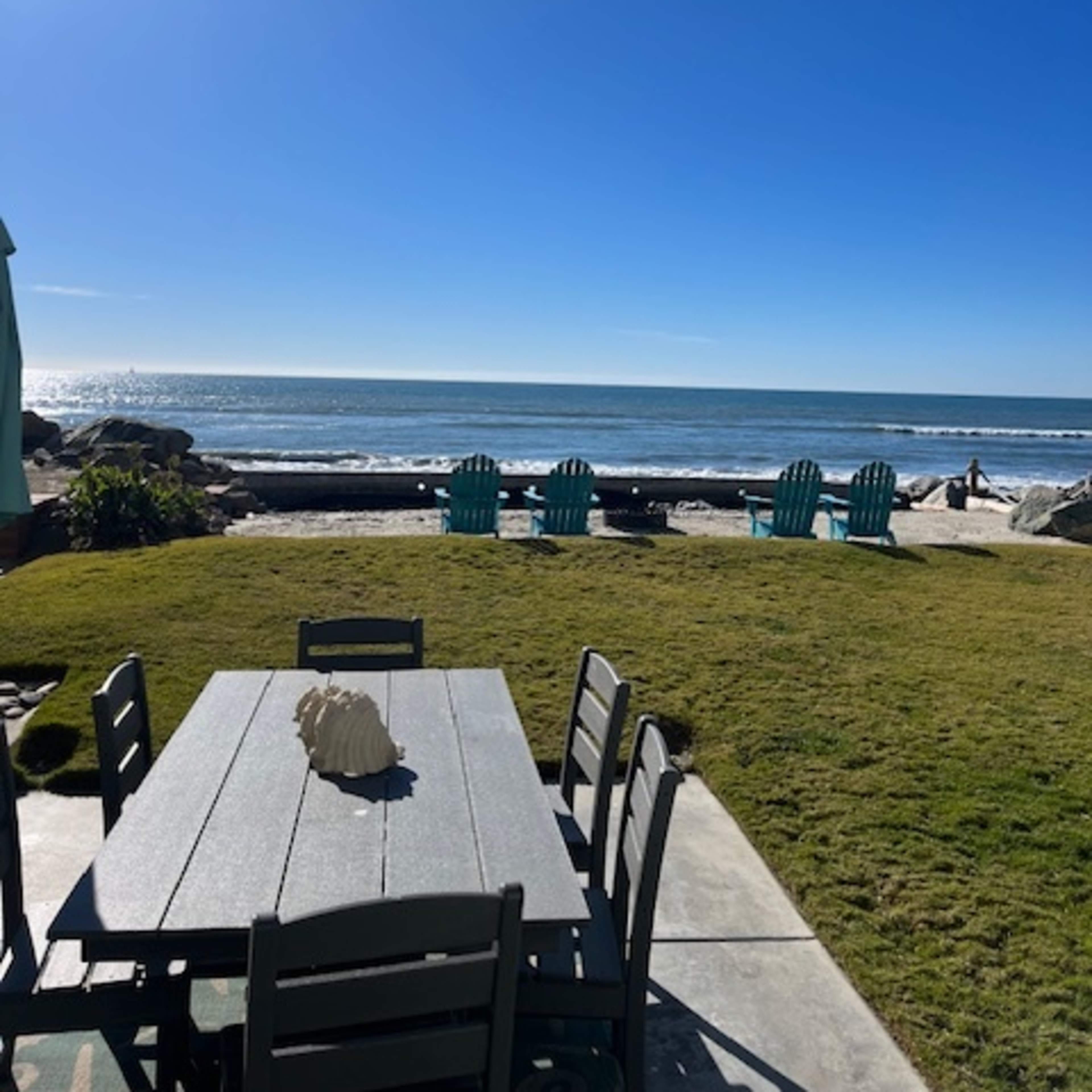 A dining table and chairs are set on a grassy area overlooking the ocean and a clear sky.
