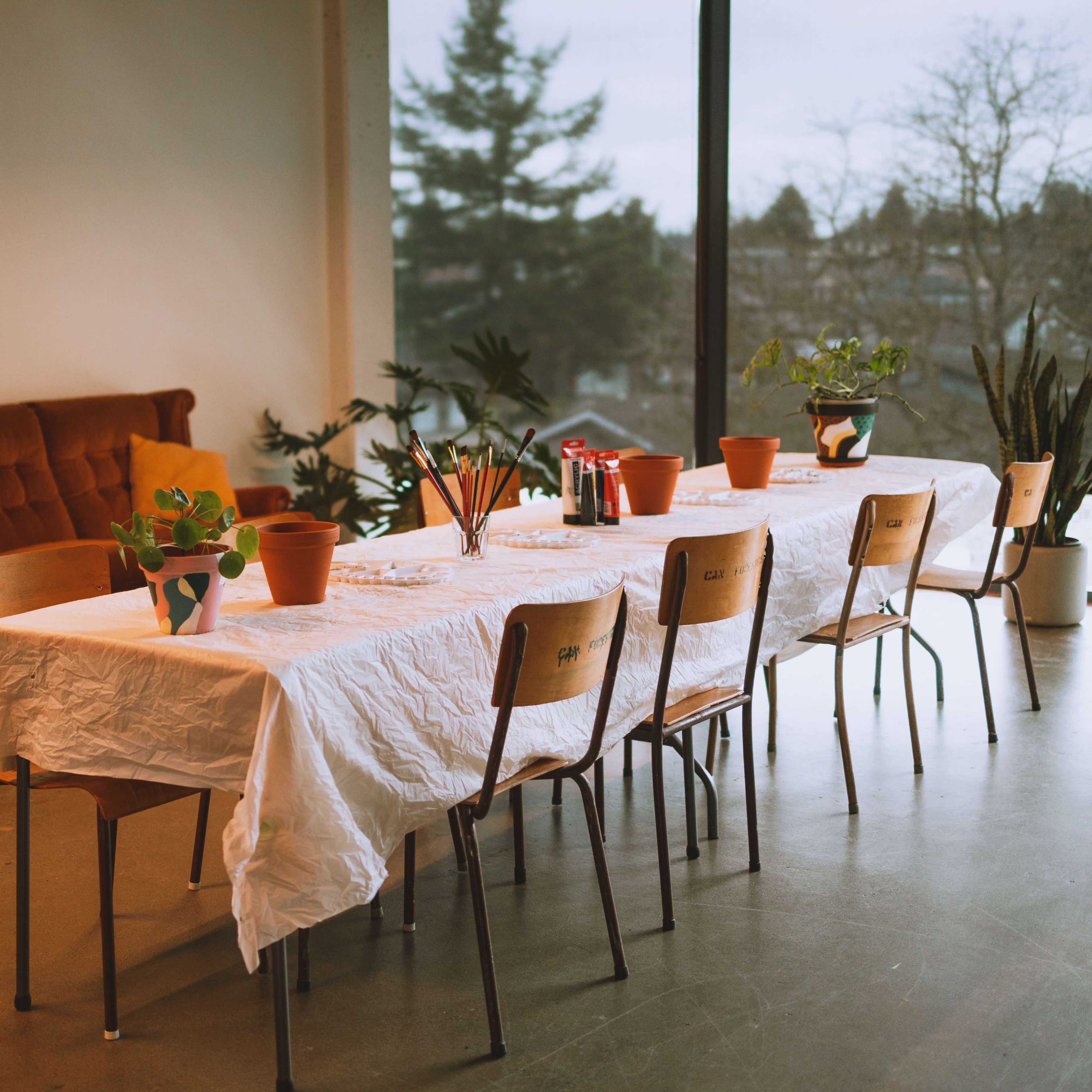 A long table covered with a white cloth is set up in a room with a sofa and potted plants, overlooking a window.