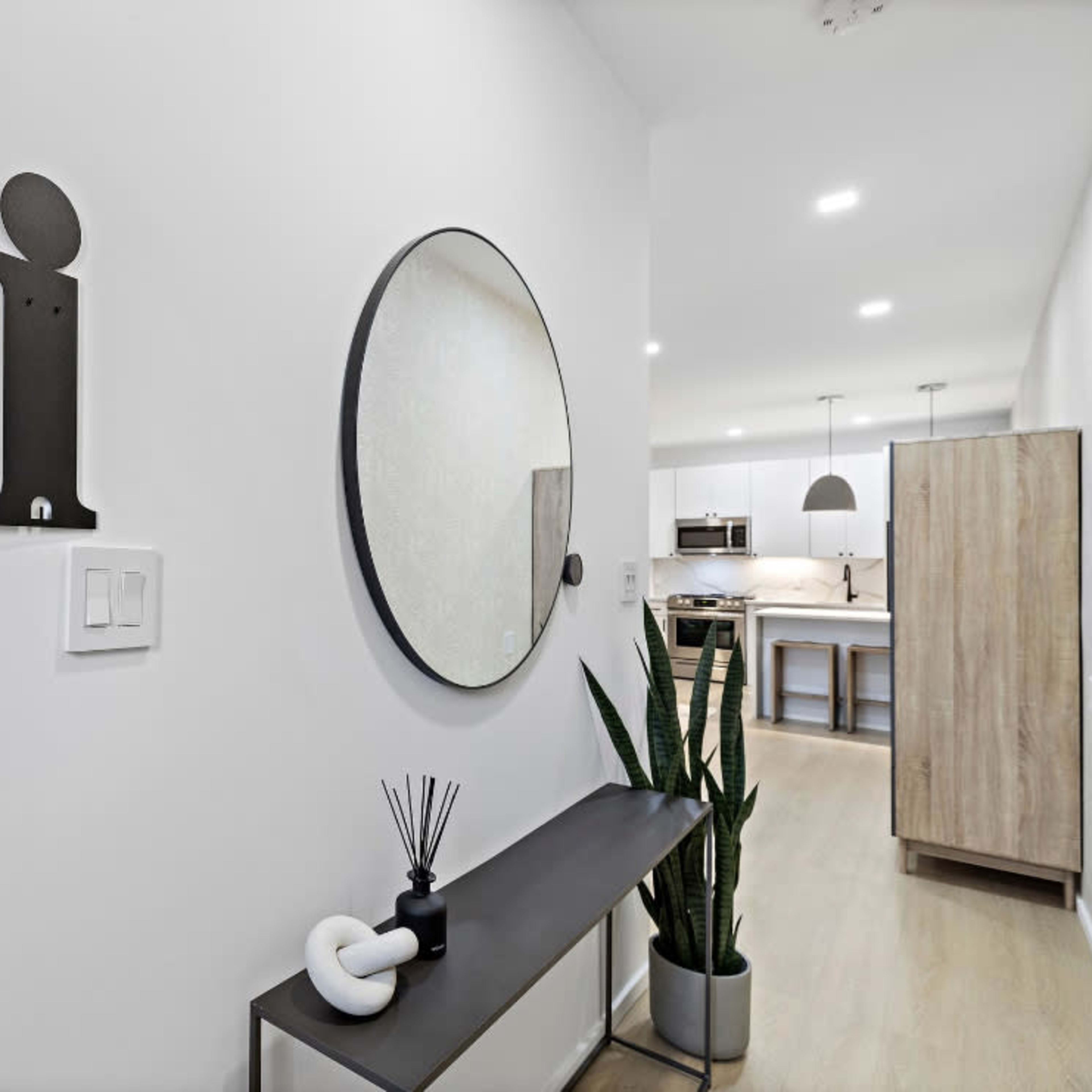 An entryway with a wall-mounted "hi" sign, a round mirror, a wooden console table with a plant, and a view of a kitchen in the background.