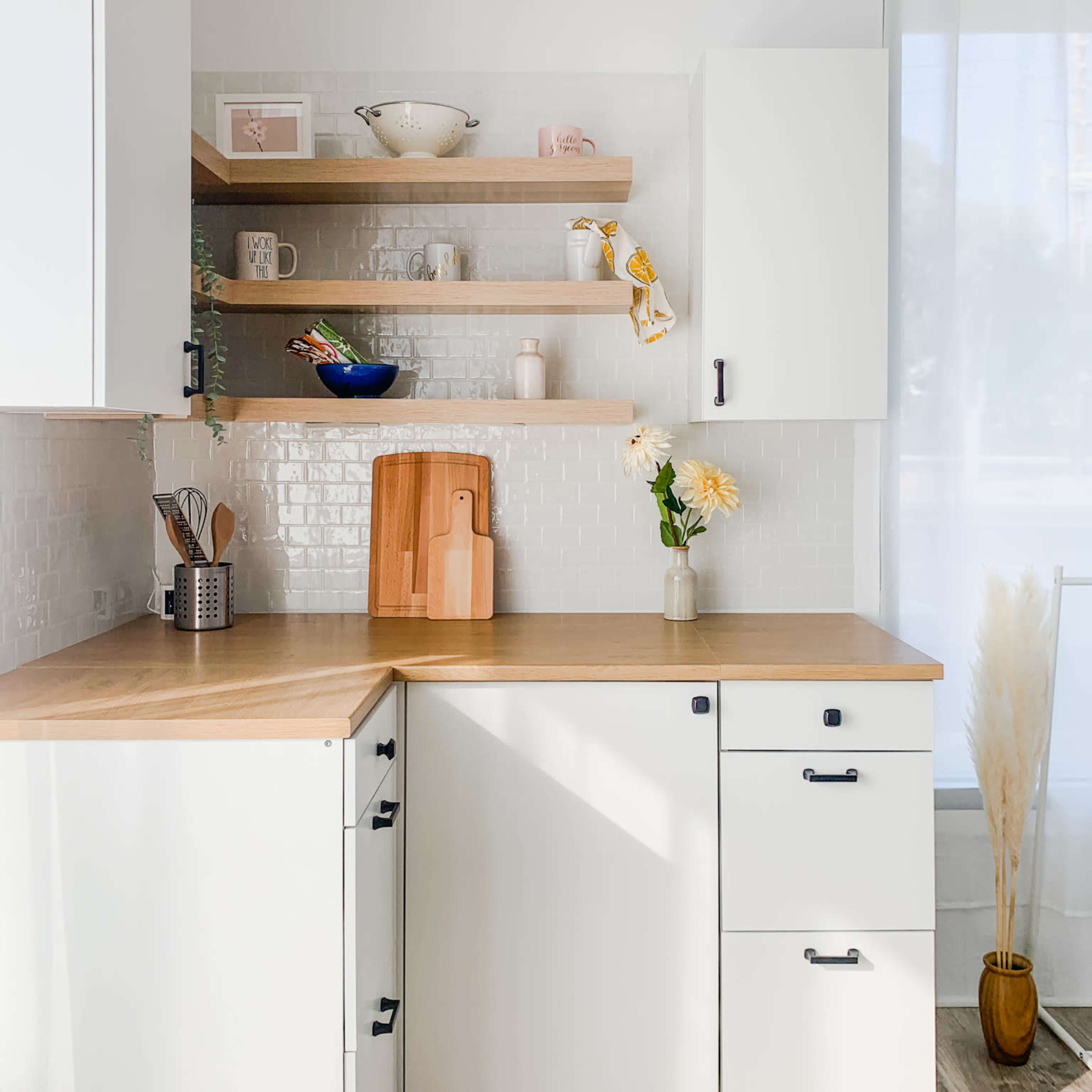 A bright kitchen features white cabinetry, wooden shelves, and a wooden countertop, with decorative items and a vase of flowers on display.