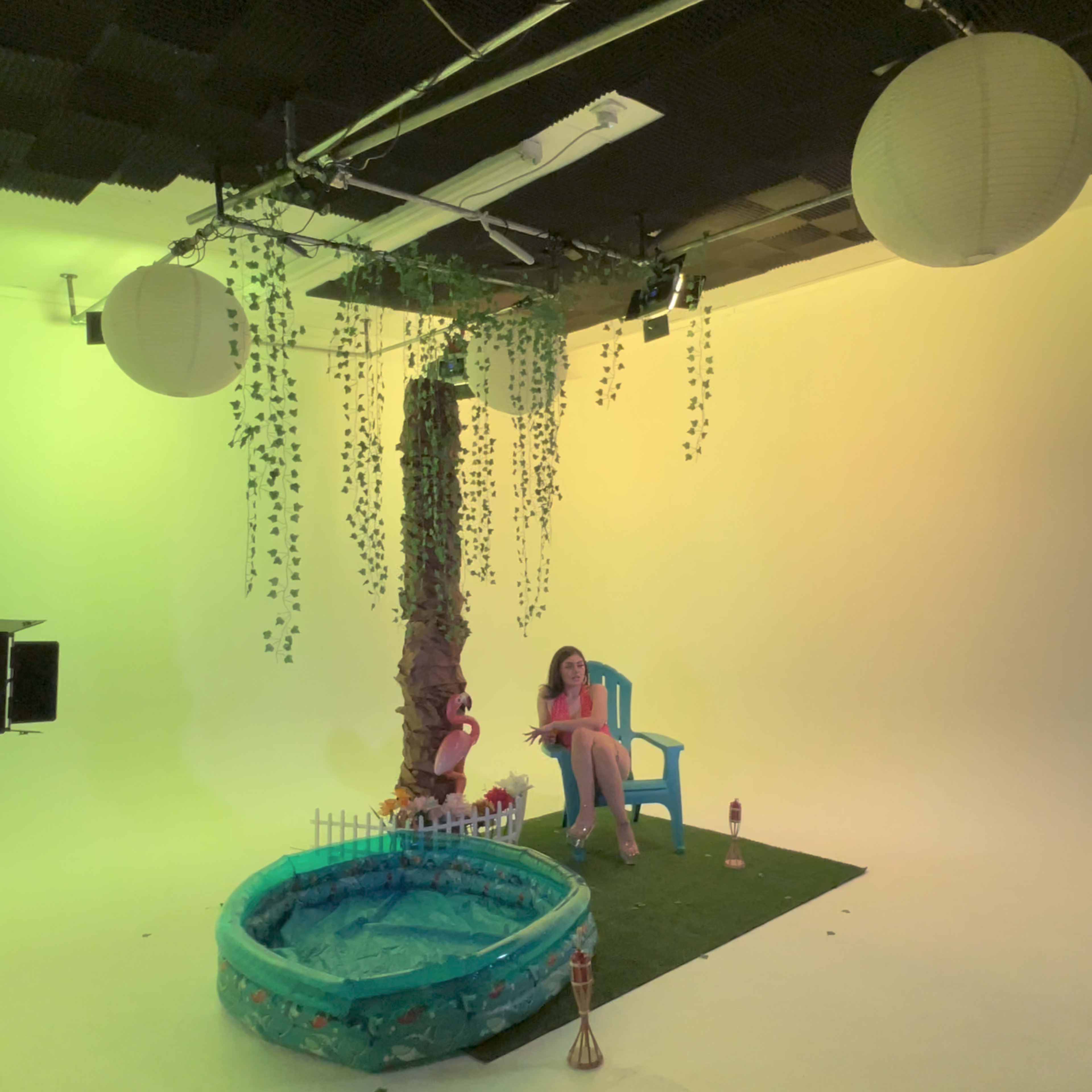 A woman sits in a blue chair beside a small inflatable pool under a large artificial tree adorned with hanging greenery in a brightly lit studio backdrop.