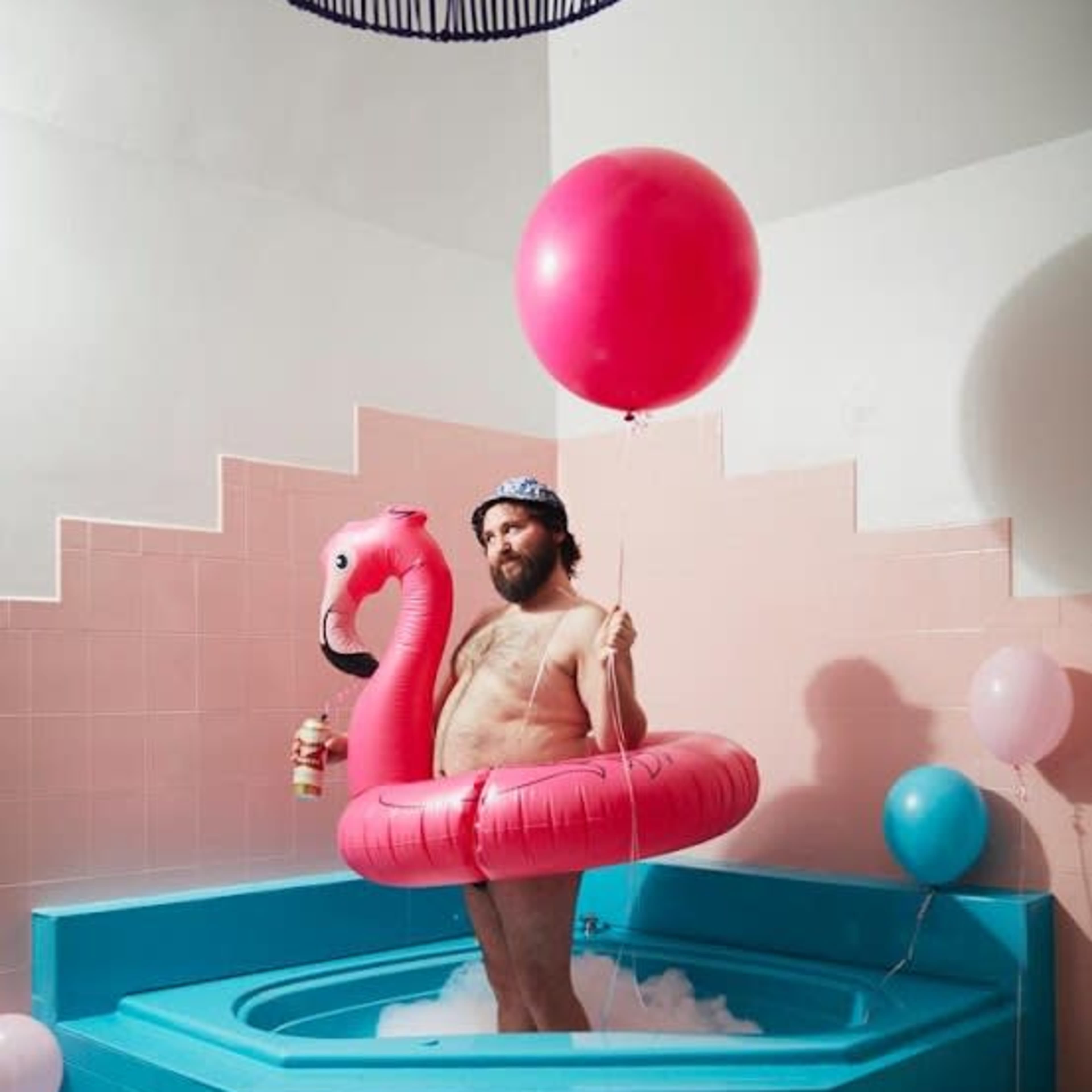A man stands in a blue bathtub wearing a pink flamingo inflatable ring, holding a drink and a balloon, surrounded by pastel-colored balloons.