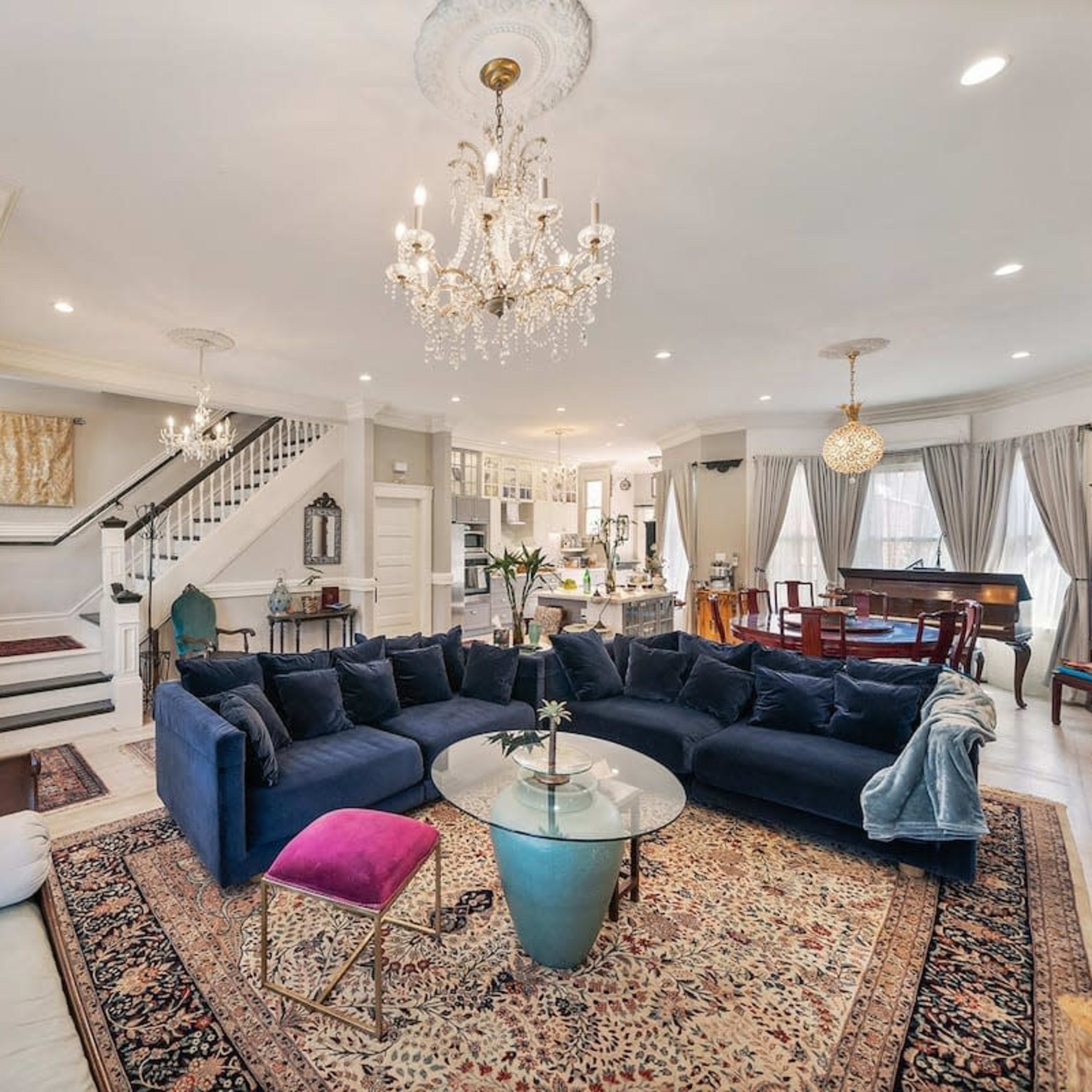 A well-lit living room with a large navy blue sectional couch, a glass coffee table, a chandelier, and a staircase leading to the upper level.
