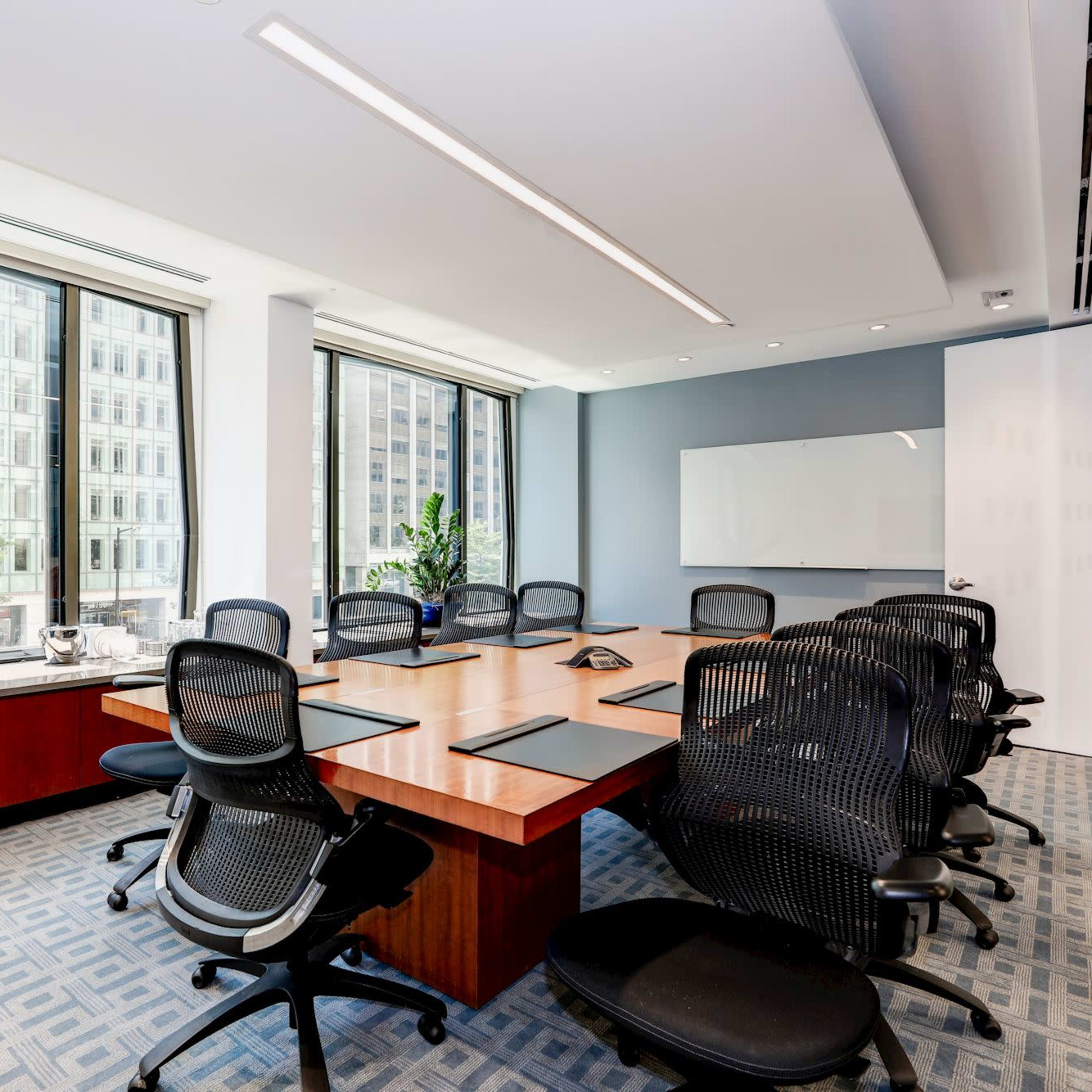 A modern conference room features a large wooden table surrounded by black office chairs, with floor-to-ceiling windows offering a view of a cityscape.
