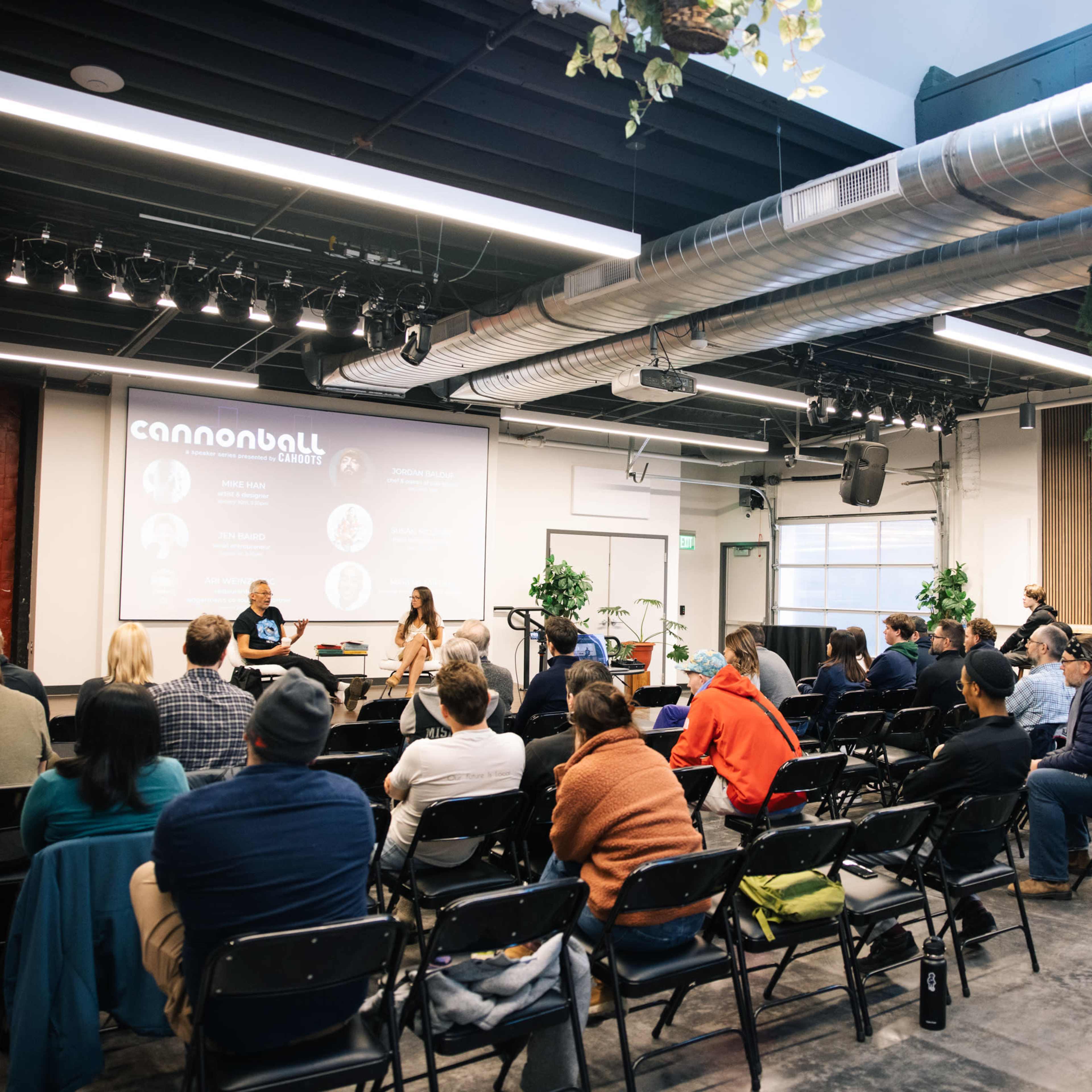 A panel discussion takes place in a spacious room filled with seated attendees, featuring speakers at a table in front of a projected screen.