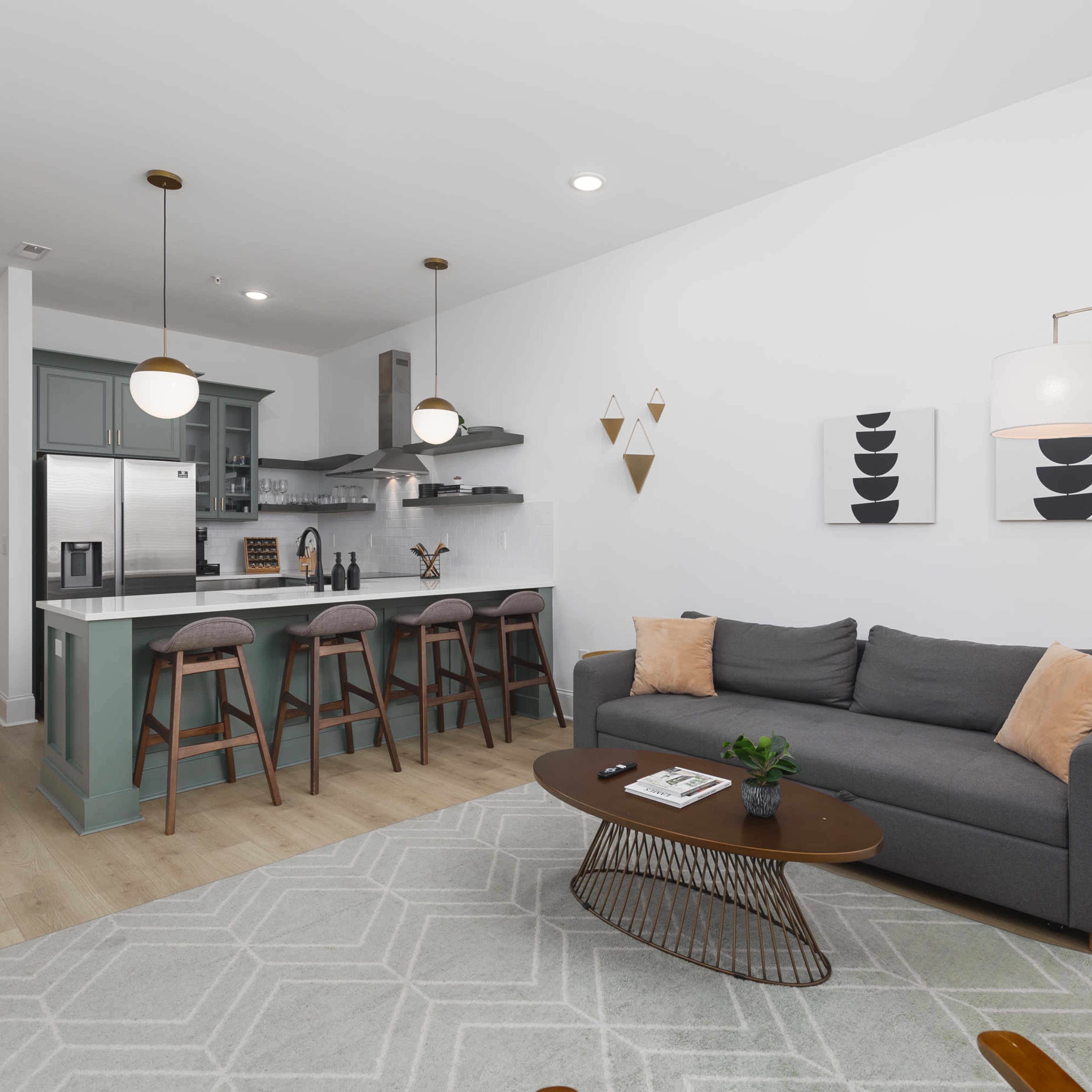 A modern living room with a gray couch, a wooden coffee table, and a kitchen area featuring bar stools and green cabinetry.