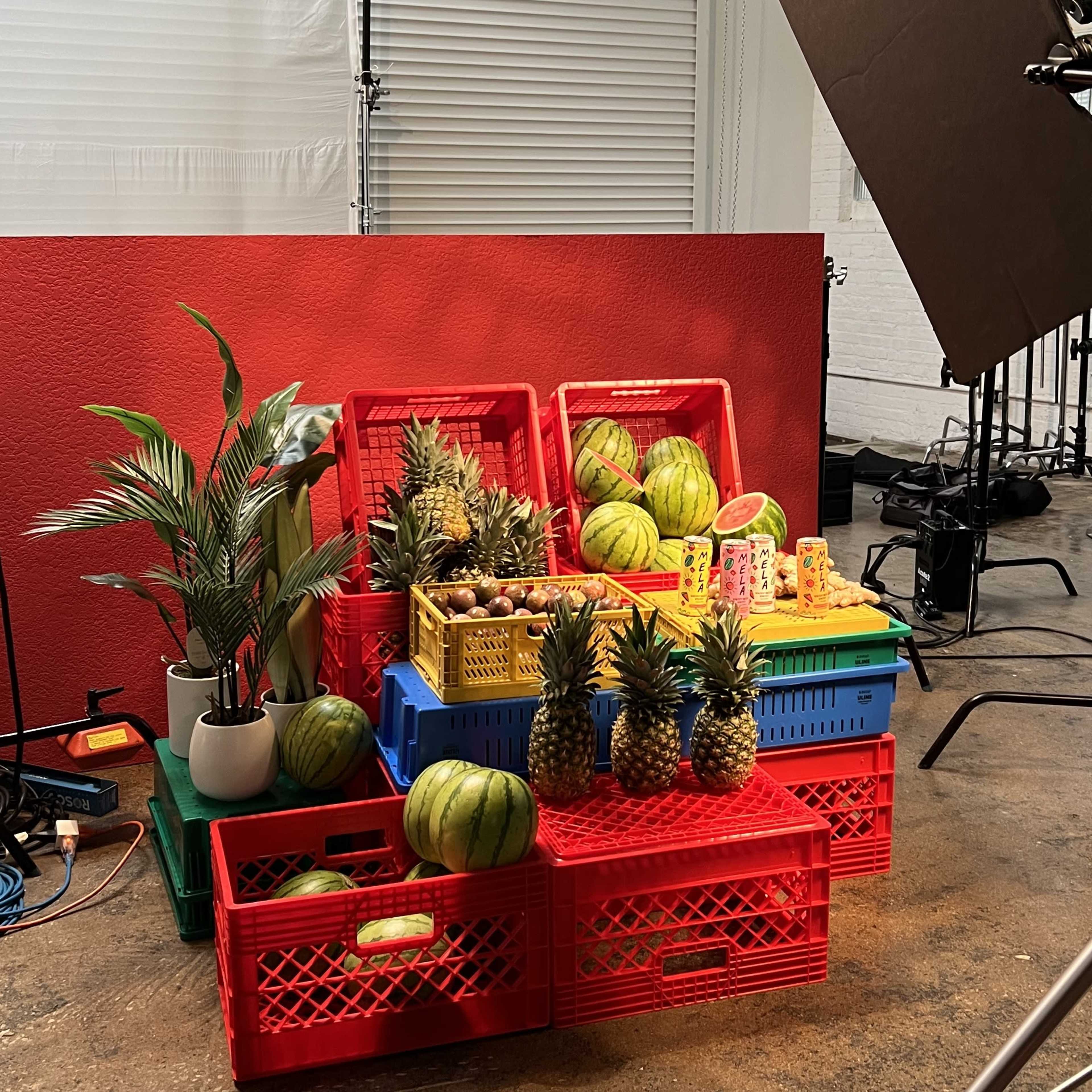 The scene features colorful crates stacked with various fruits, including watermelons and pineapples, arranged beside a potted plant, in a studio setting.