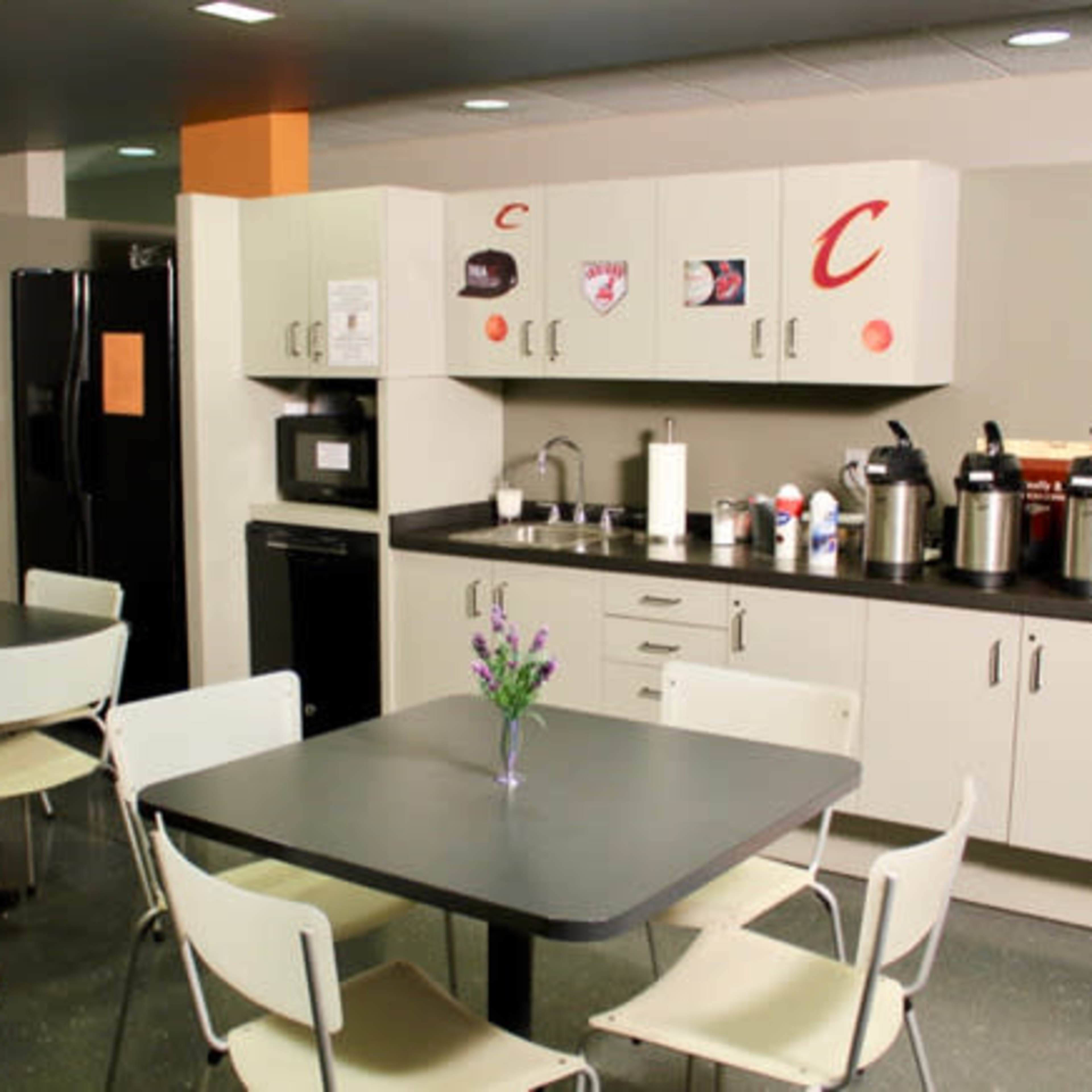 A kitchen area features a countertop with coffee machines, a microwave, and a sink, along with tables and chairs arranged for dining.