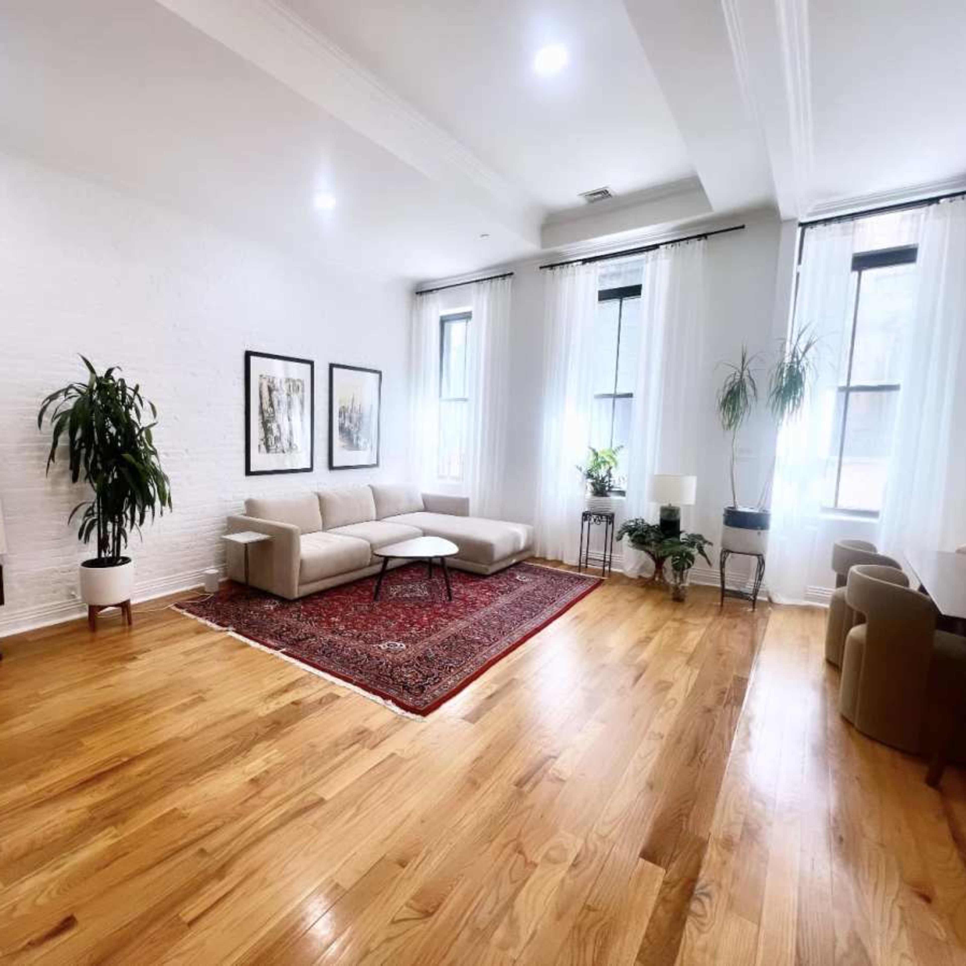 A spacious living room featuring a light-colored sofa, a round coffee table, a rug, and large windows adorned with sheer curtains.