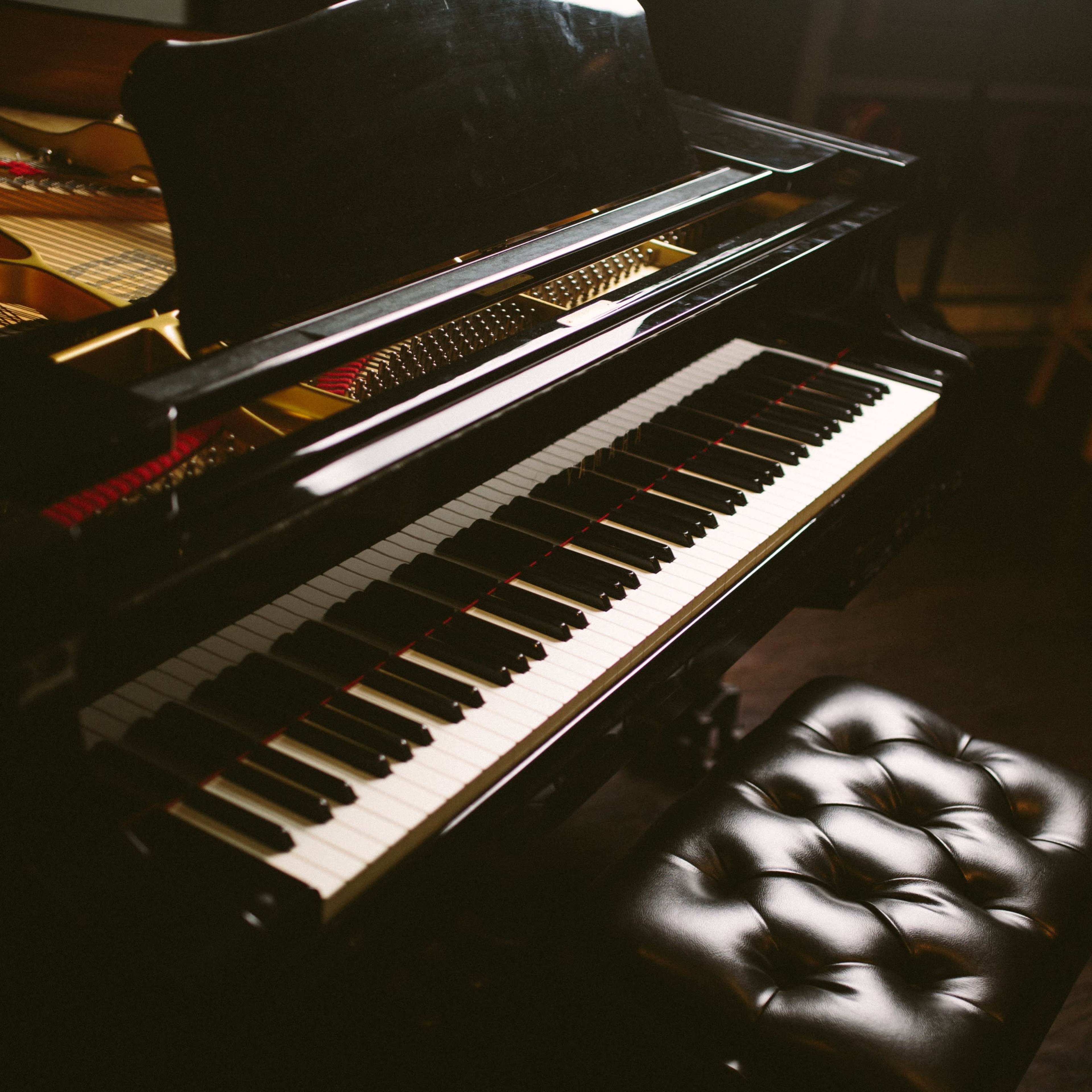 A black grand piano is positioned next to a wooden stool, with its polished keys visible in a dimly lit room.