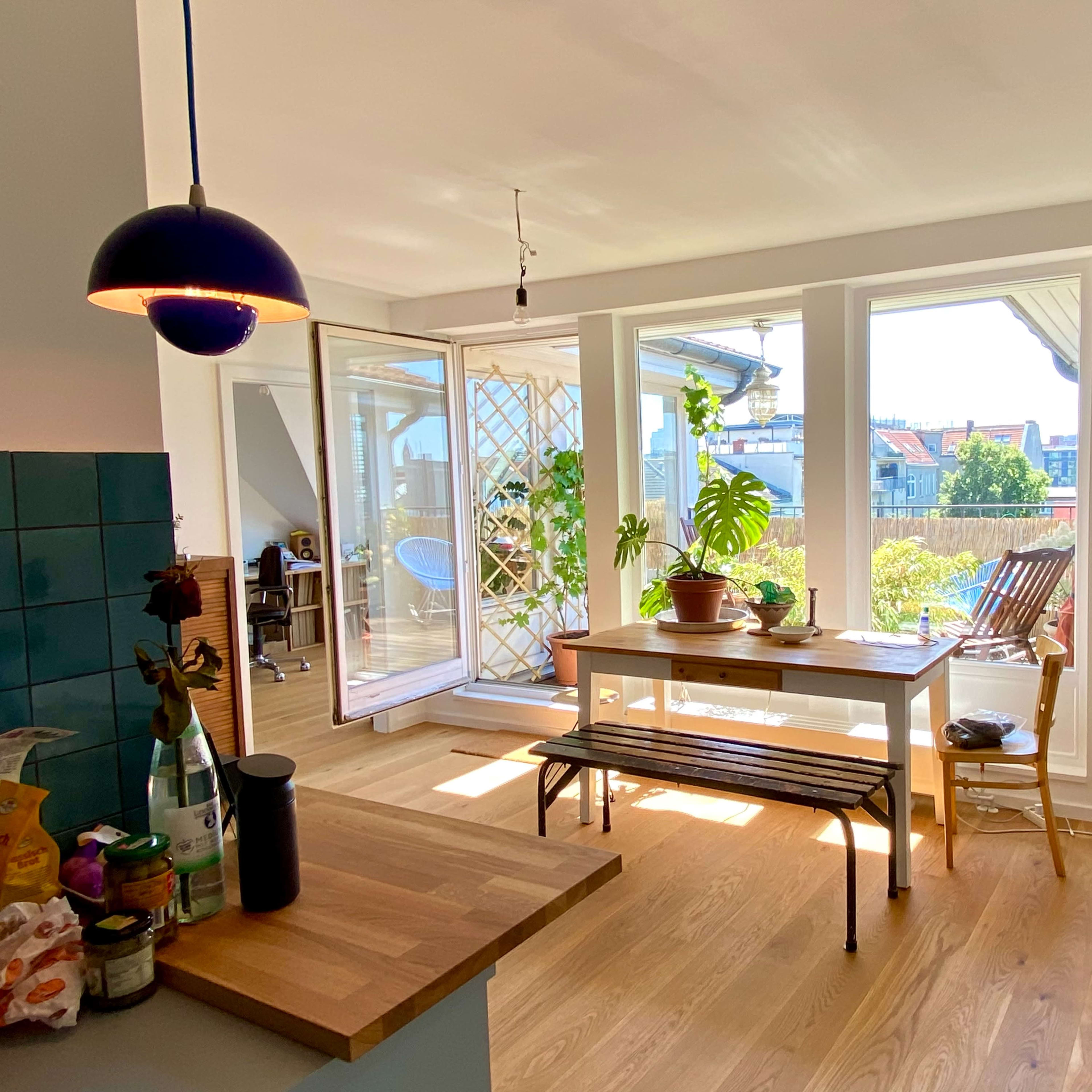 A bright, open kitchen and dining area features a wooden table surrounded by plants and large windows overlooking a balcony.