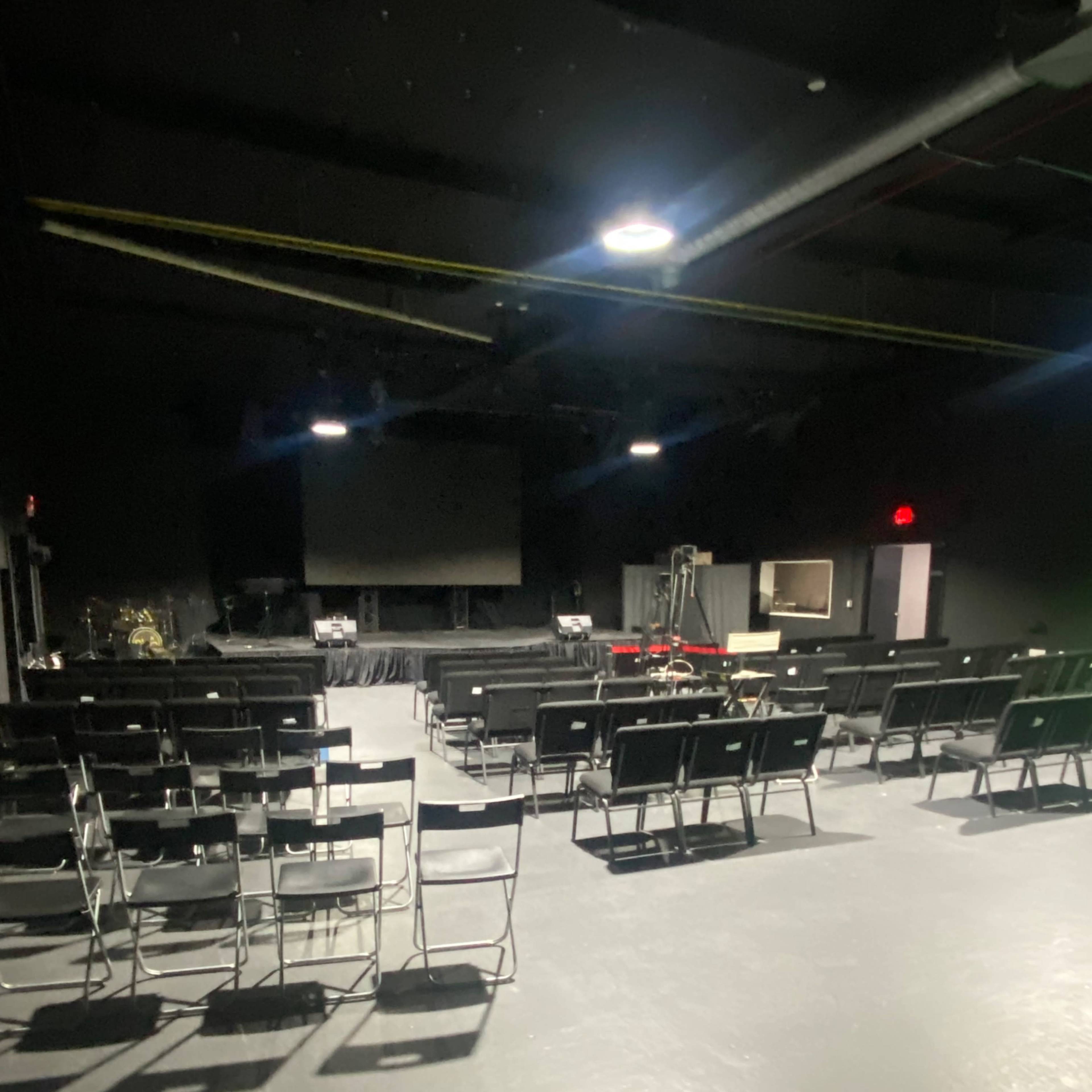 An empty indoor auditorium with rows of black chairs facing a large screen at the front.