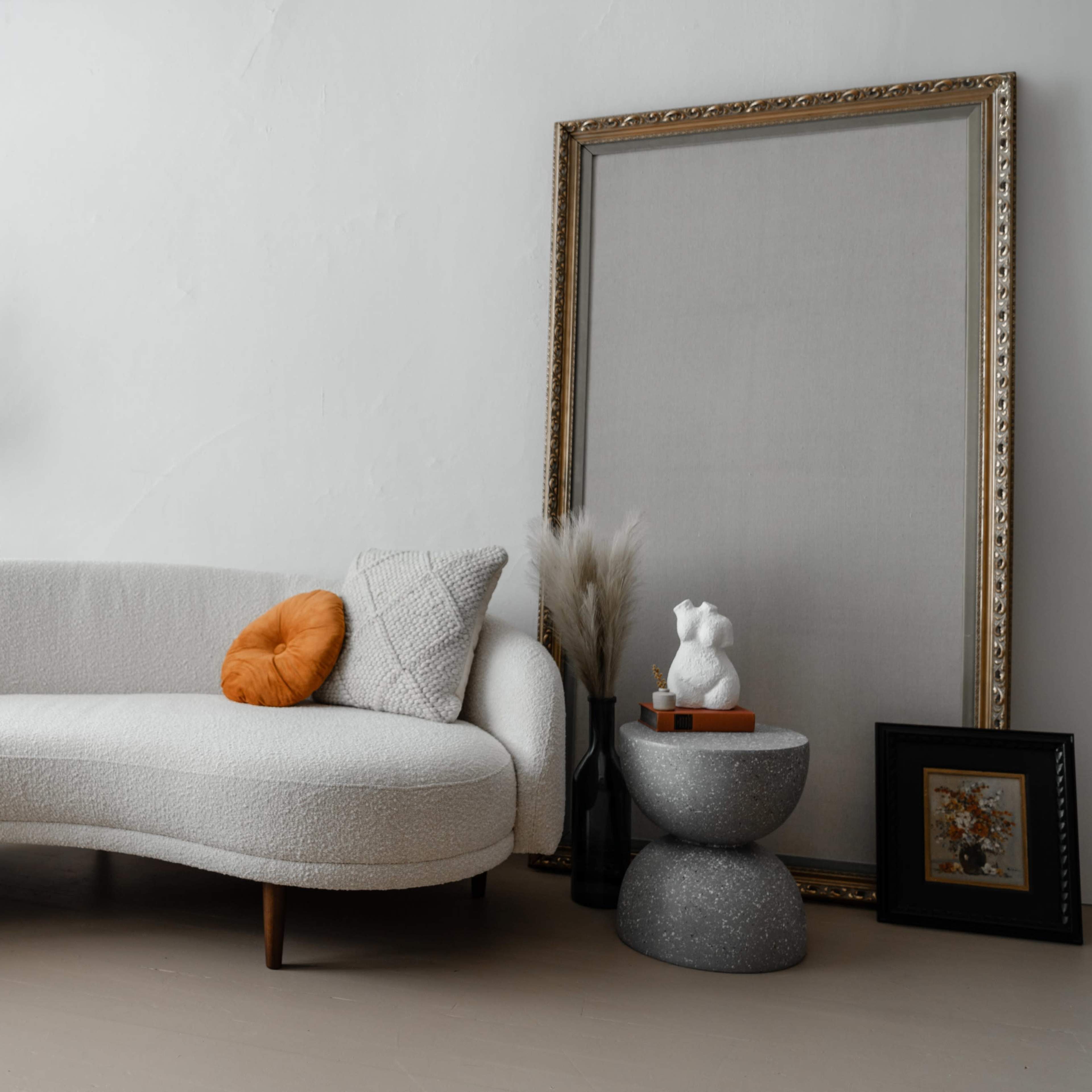 The scene features a light-colored, curved sofa with an orange cushion, accompanied by decorative elements including a potted plant, a large framed artwork, and a small side table with sculptures and framed pictures.