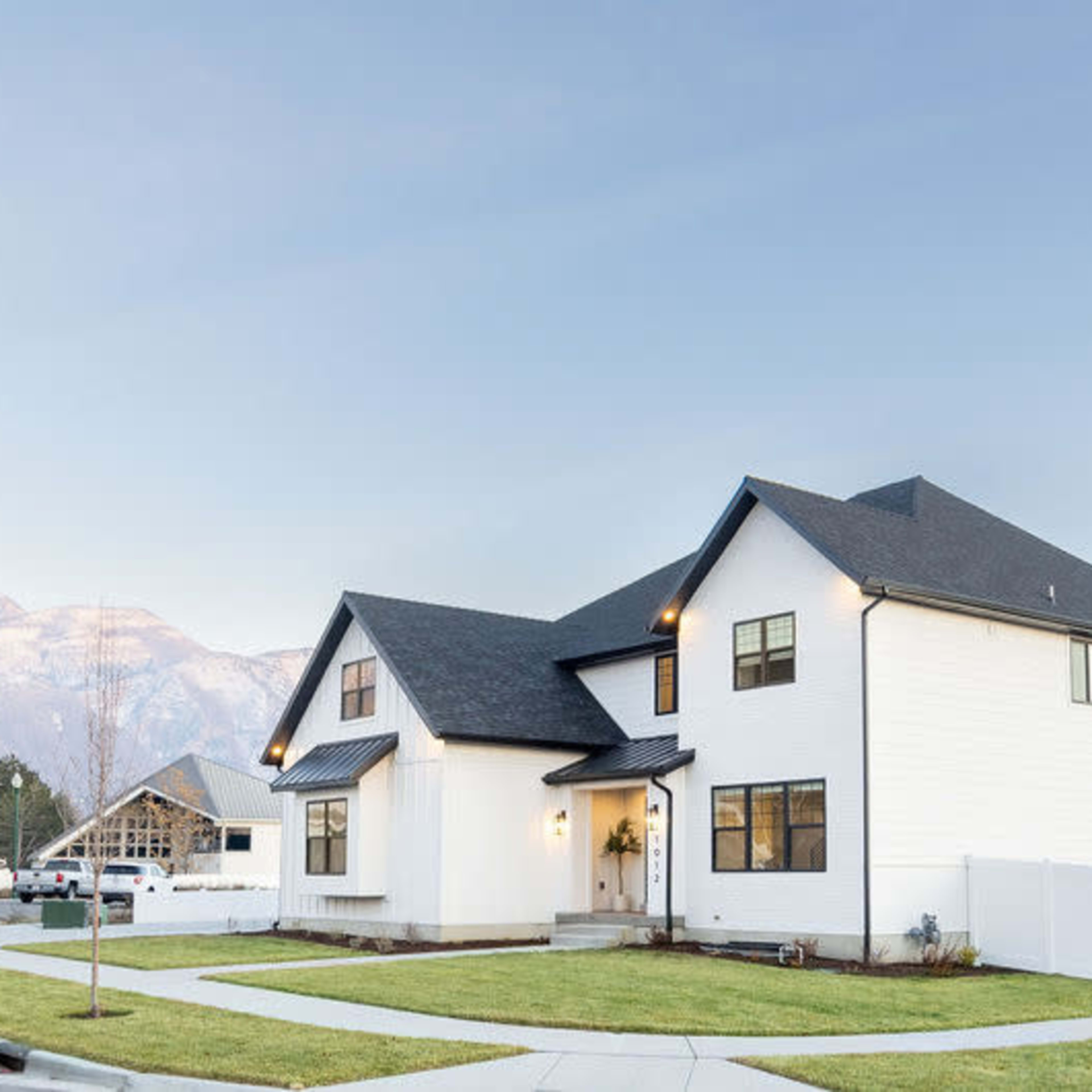 A two-story white house is situated on a corner lot with a manicured lawn, surrounded by a white fence and mountains in the background.