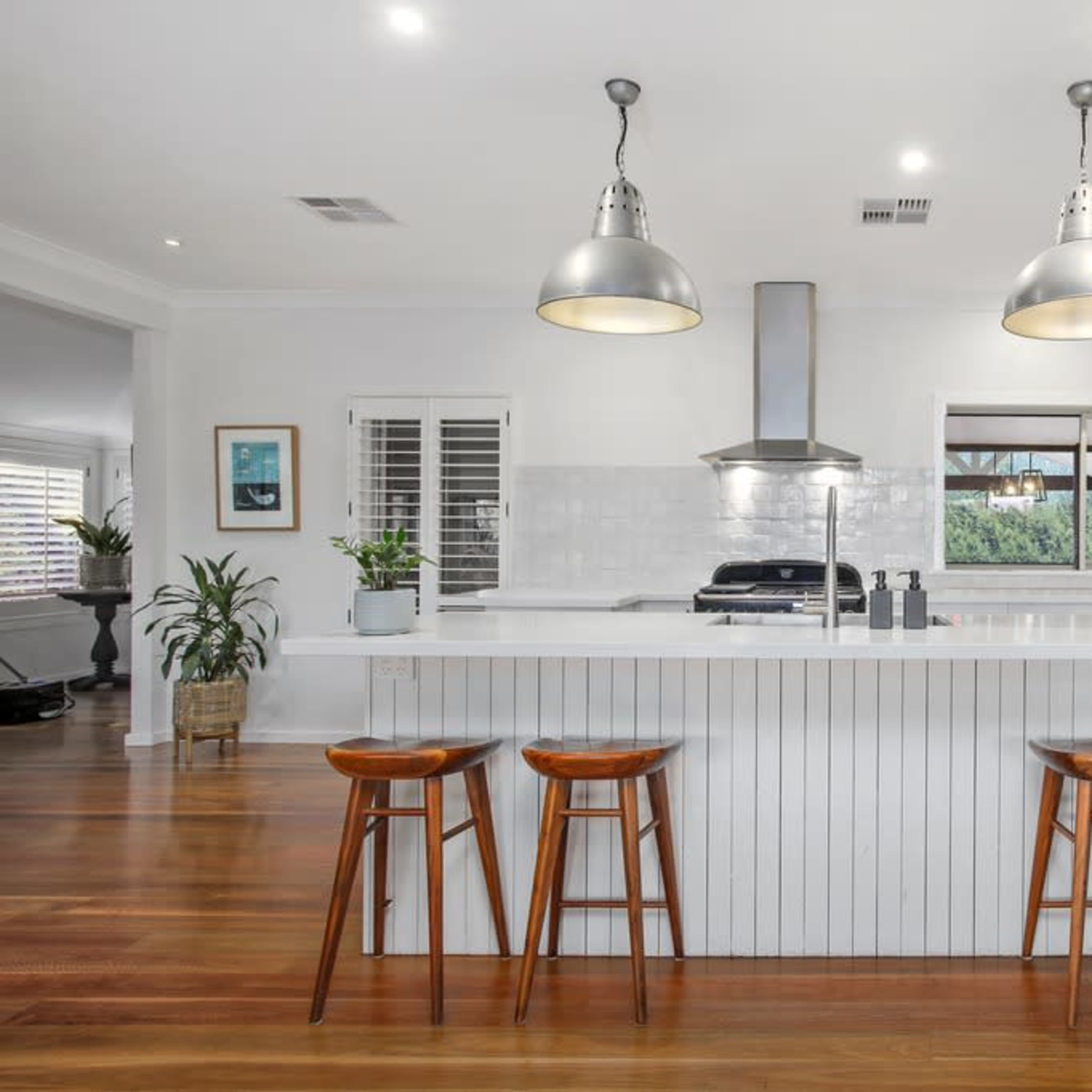 A modern kitchen features a white island with three wooden stools, pendant lighting, and stainless steel appliances.
