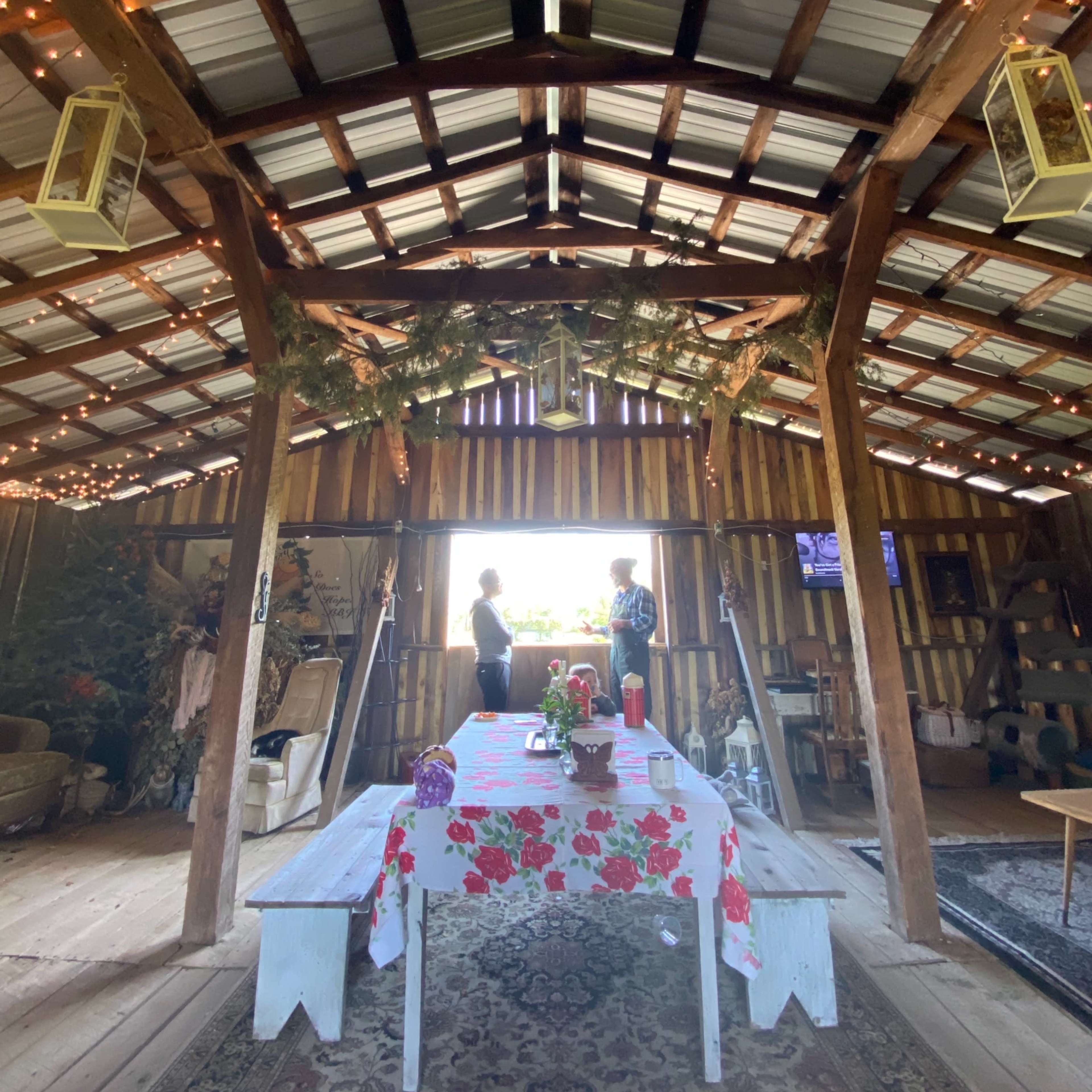 The interior of a rustic barn features a table with a floral tablecloth and two people standing by a window, surrounded by wooden beams and decorative lights.