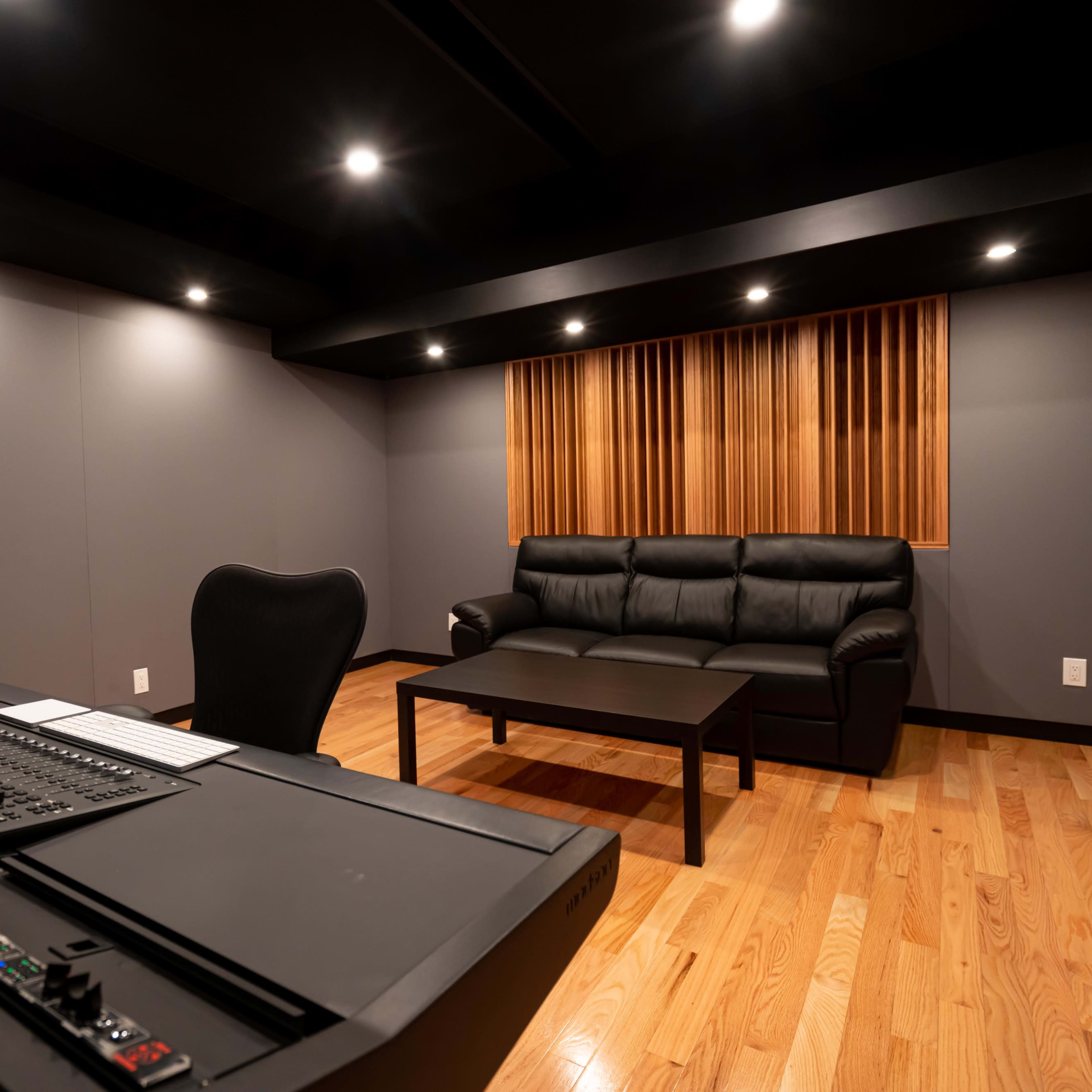 A modern control room with a black desk equipped with audio mixing equipment, a black chair, and a leather couch positioned near a wooden coffee table against a wall with soundproofing panels.