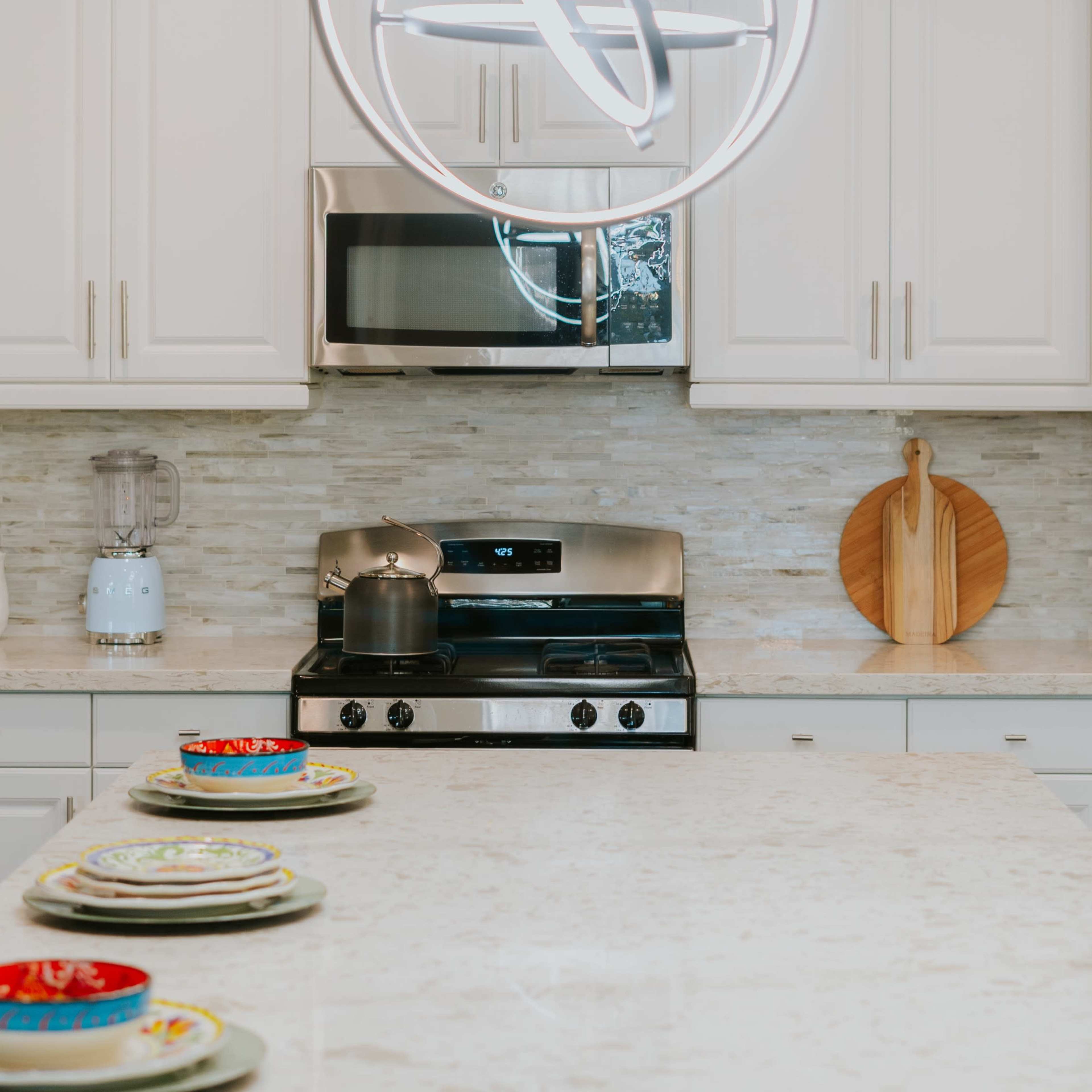A modern kitchen features white cabinets, a stainless steel stove, a circular pendant light, and a marble countertop with dishes arranged neatly.