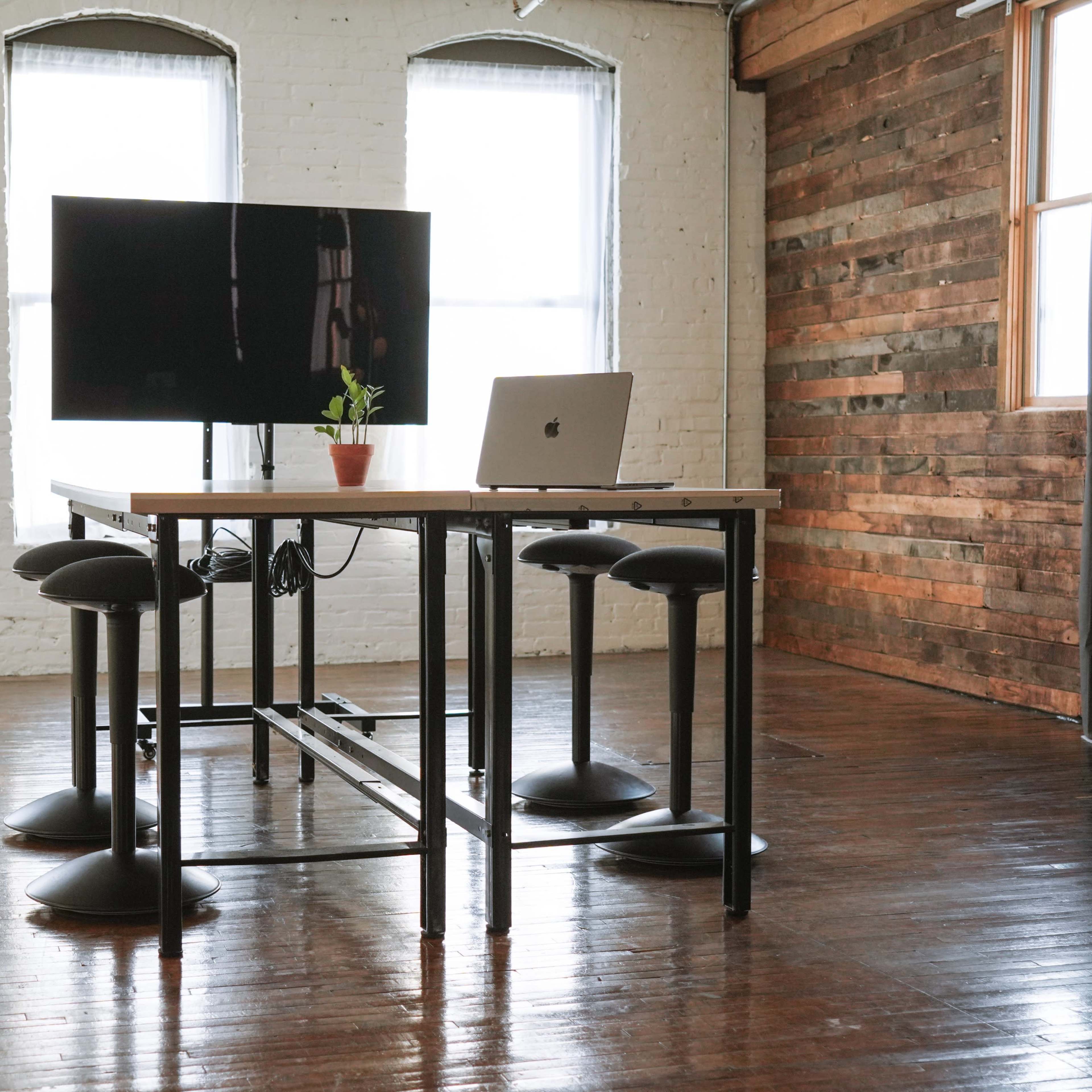 A work desk with a laptop and a potted plant stands beside a large monitor in a spacious, well-lit room with wooden walls.