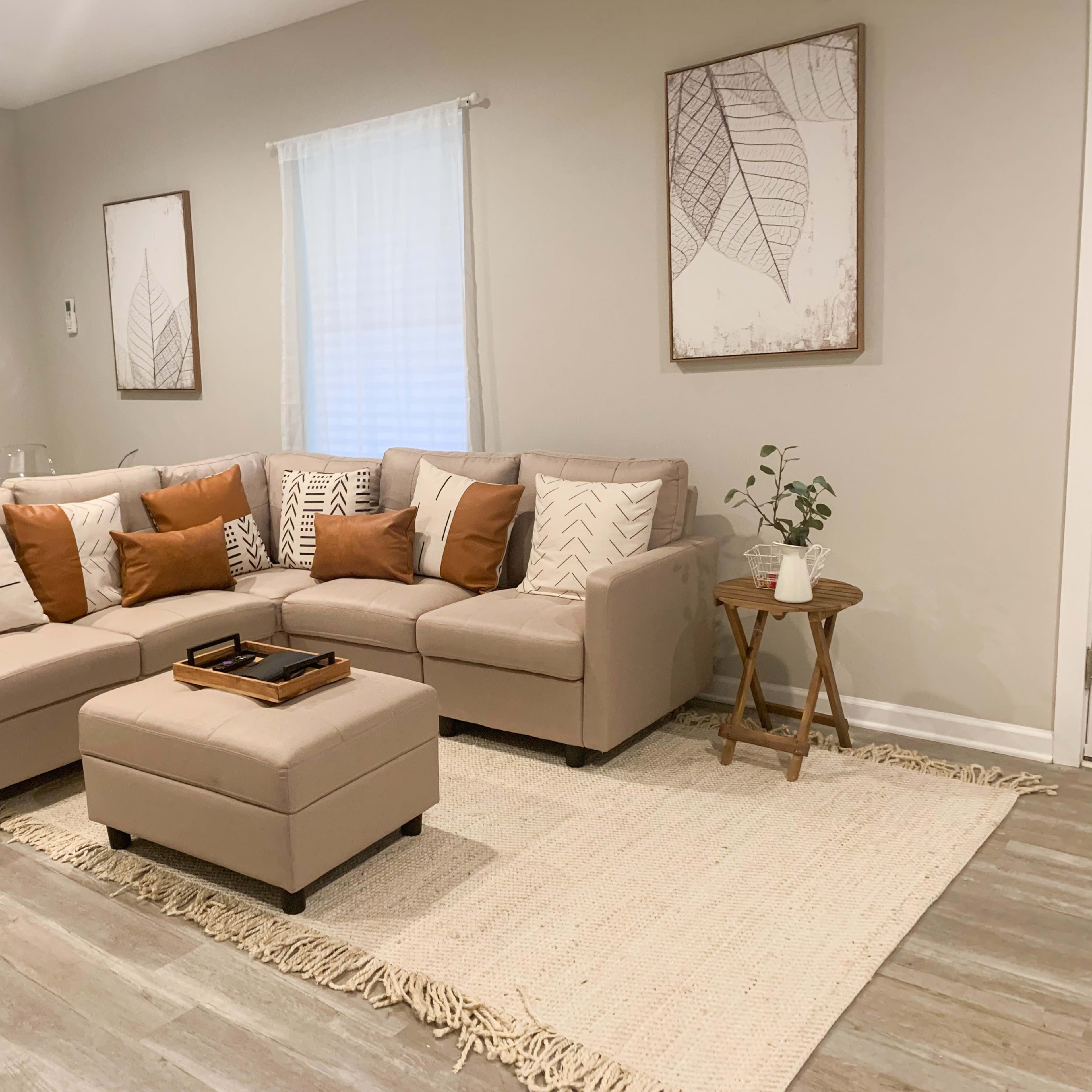 A light-colored living room featuring a sectional sofa with patterned cushions, a small ottoman, a woven area rug, and two framed leaf-themed artworks on the walls.