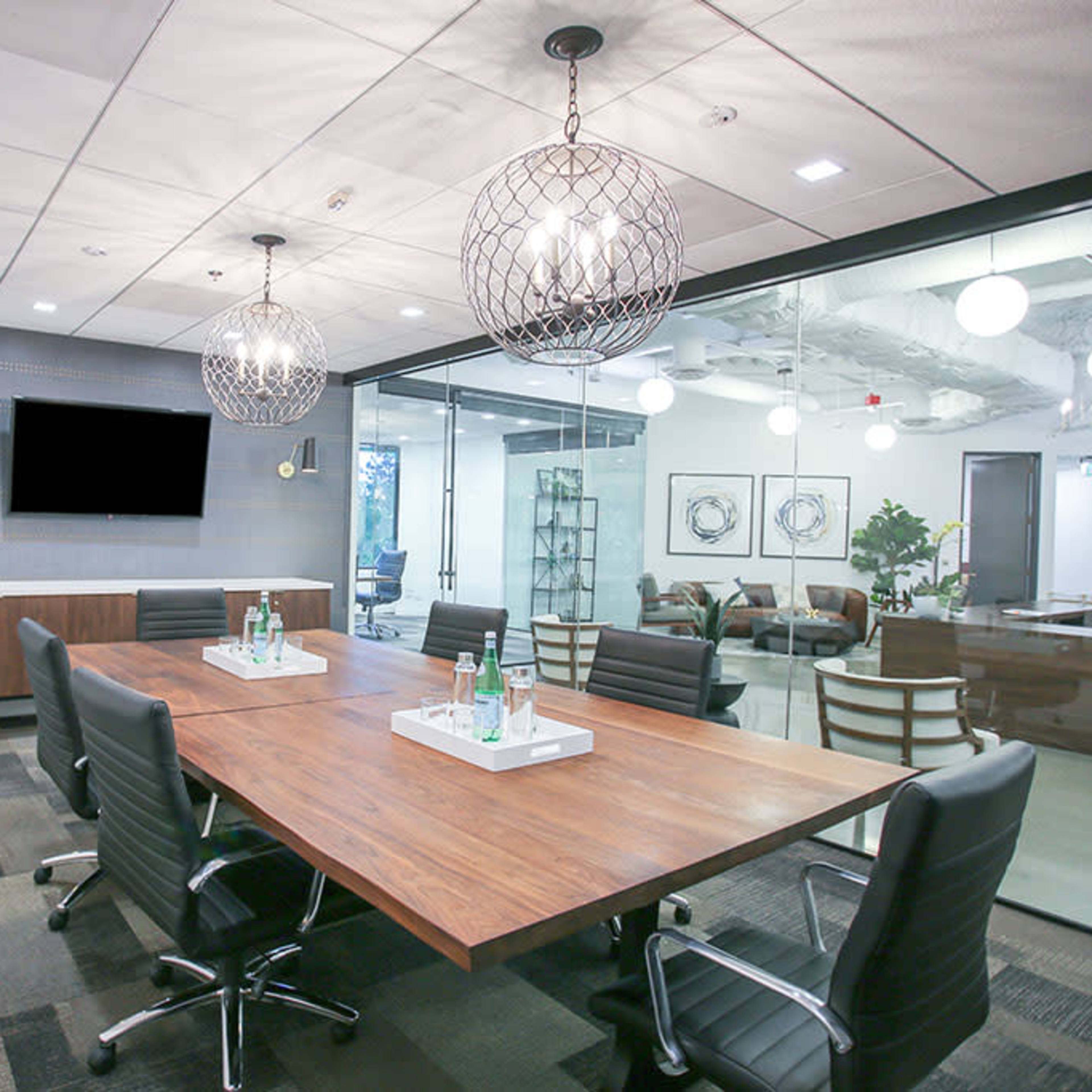 A modern conference room featuring a large wooden table surrounded by black leather chairs, with two decorative light fixtures and a flat-screen TV mounted on the wall.
