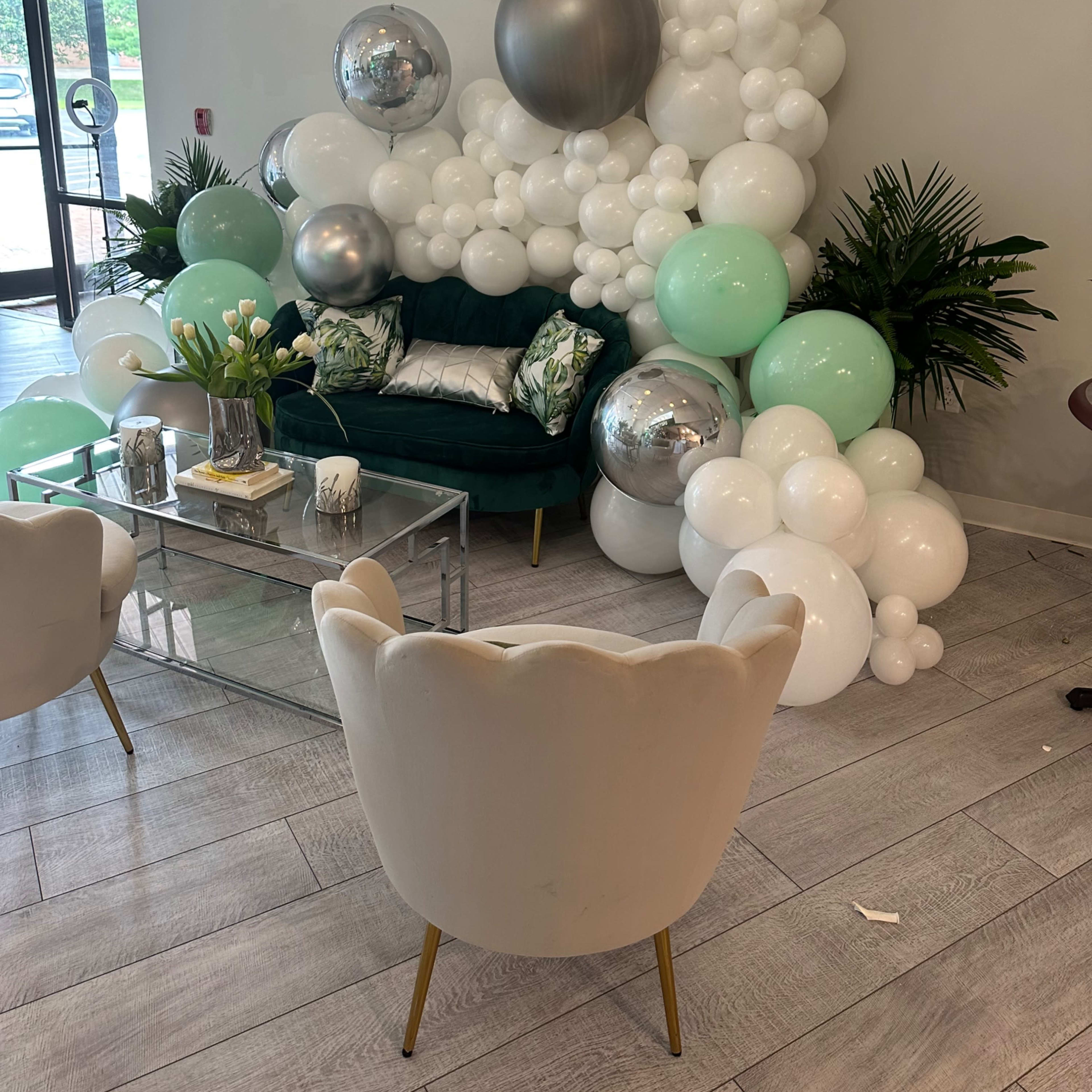 A cozy seating area with a green couch surrounded by a decorative arrangement of white and mint balloons, silver spheres, and potted plants.