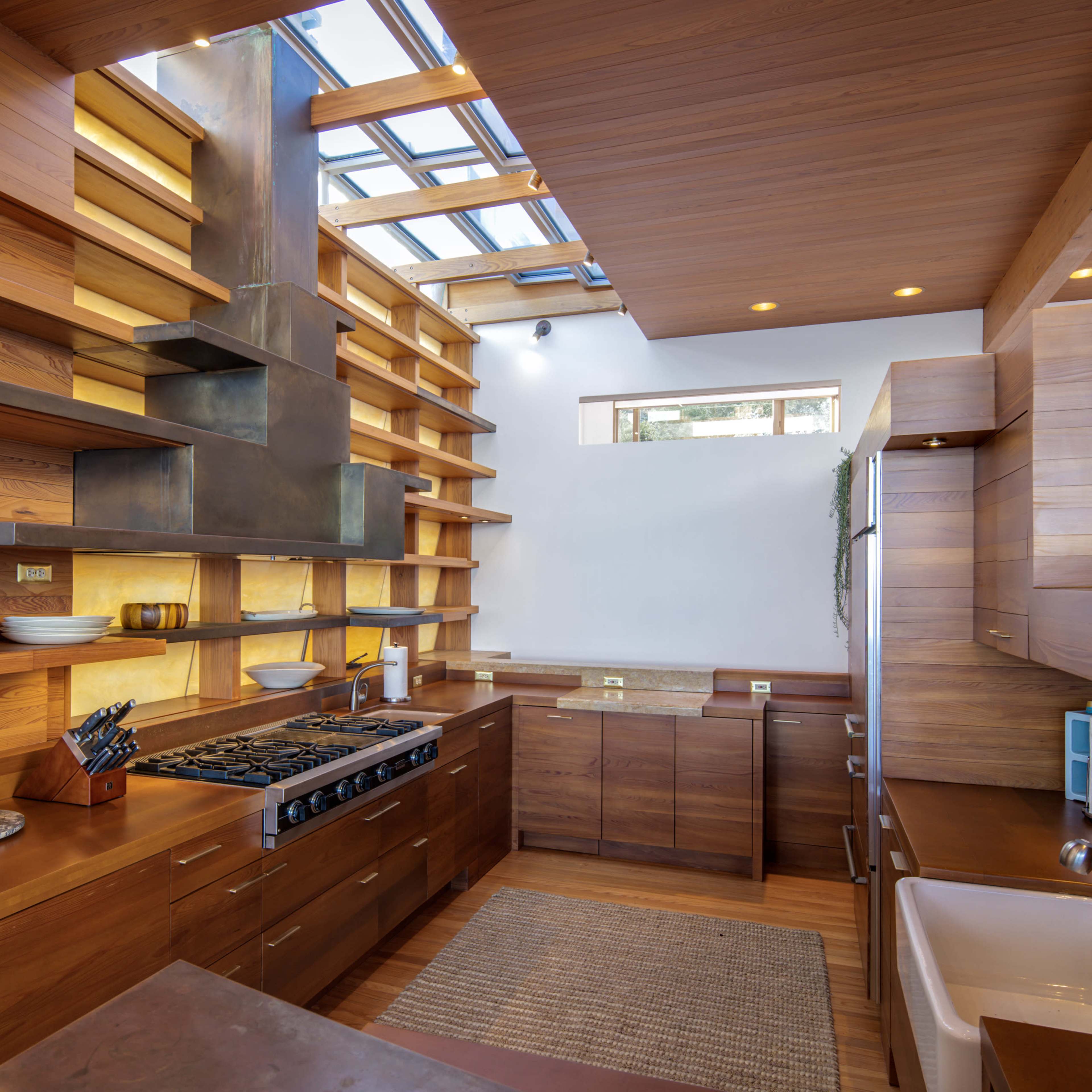 A modern kitchen with wooden cabinetry, open shelving, a stainless steel stove, and a sink under a skylight.