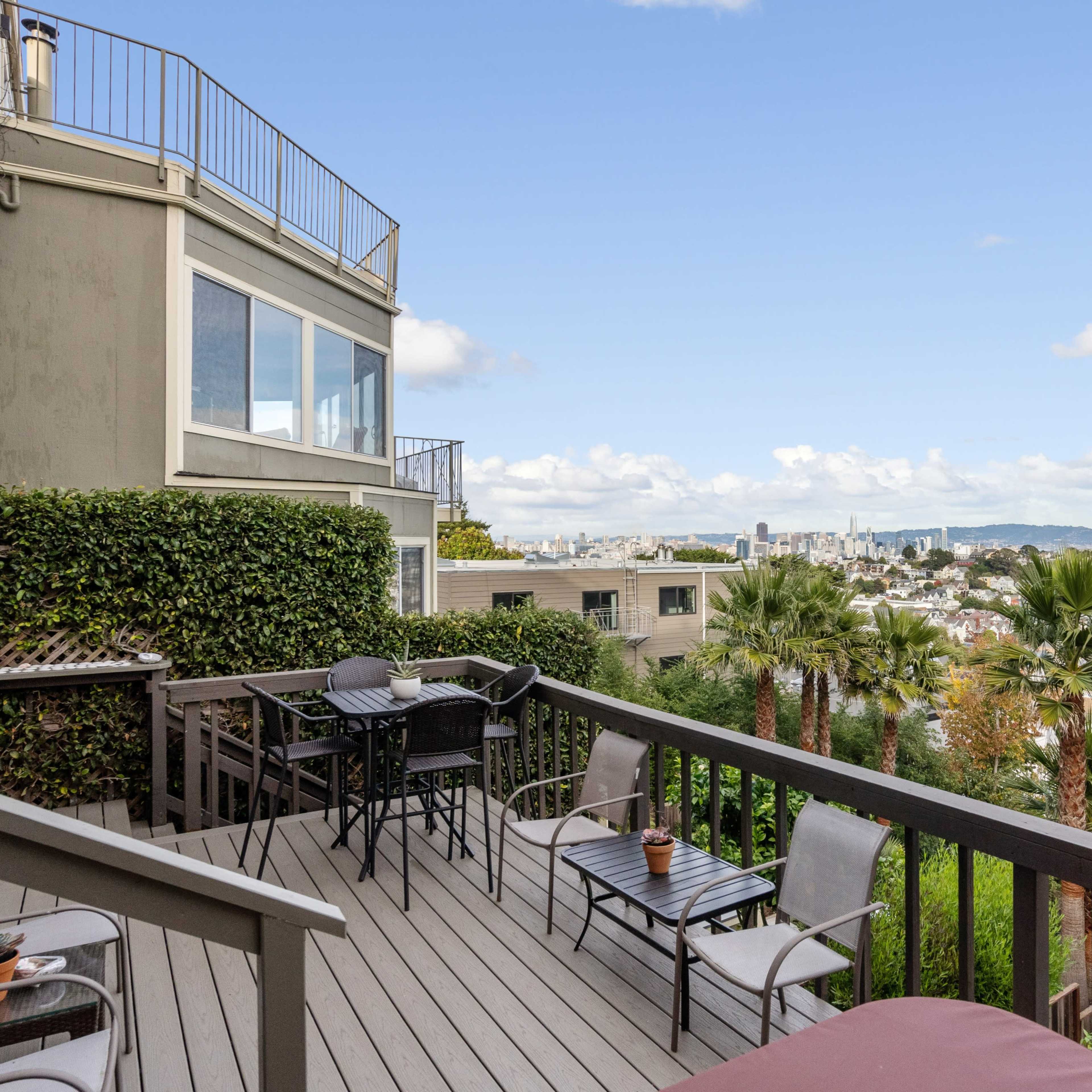 A wooden deck with seating that overlooks a cityscape, featuring palm trees and distant hills under a partly cloudy sky.