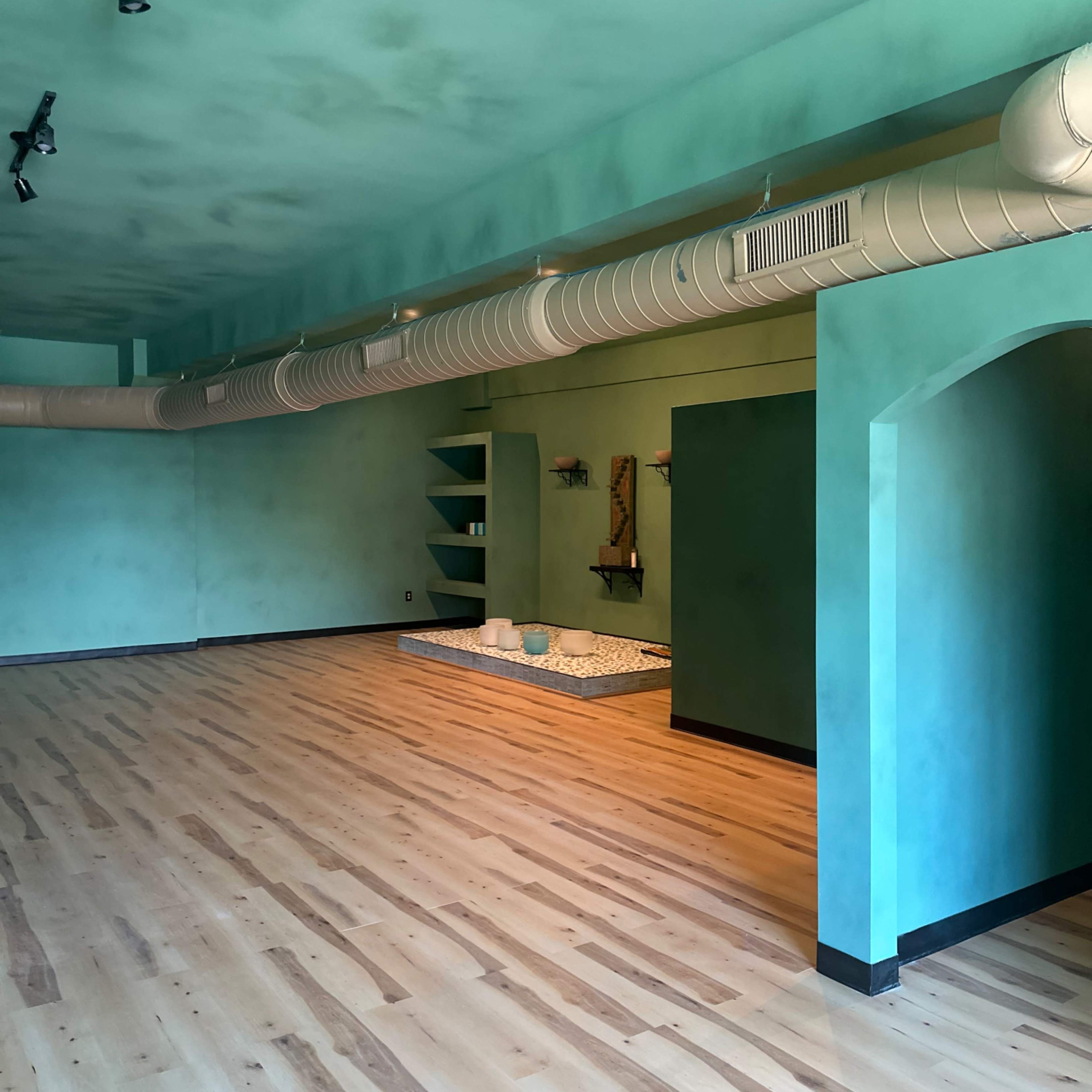 An empty room with teal walls, wooden flooring, and a central area featuring a decorative mat and a small water feature.