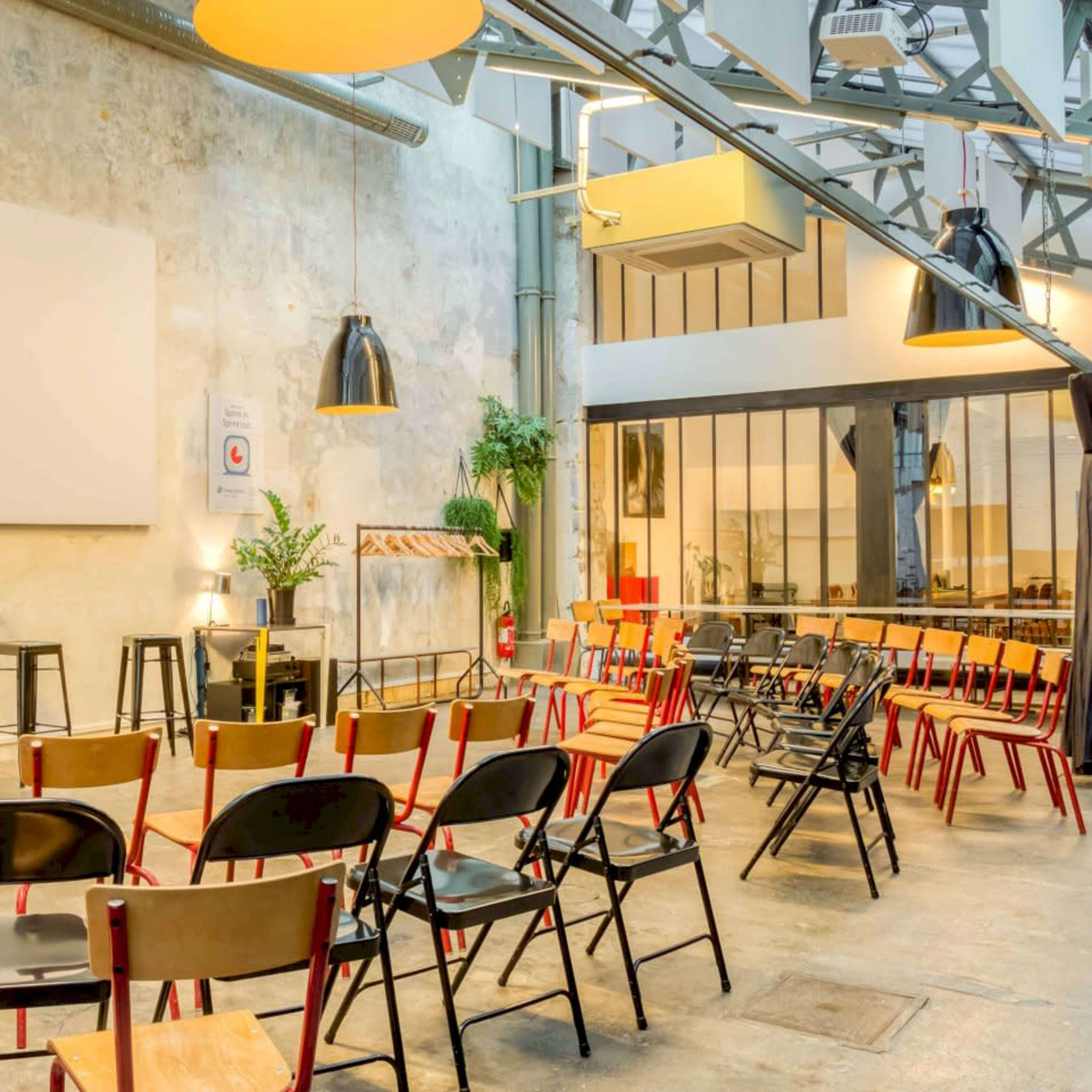 A modern event space features rows of wooden chairs arranged in front of a large screen against a textured wall, with plants and industrial lighting.