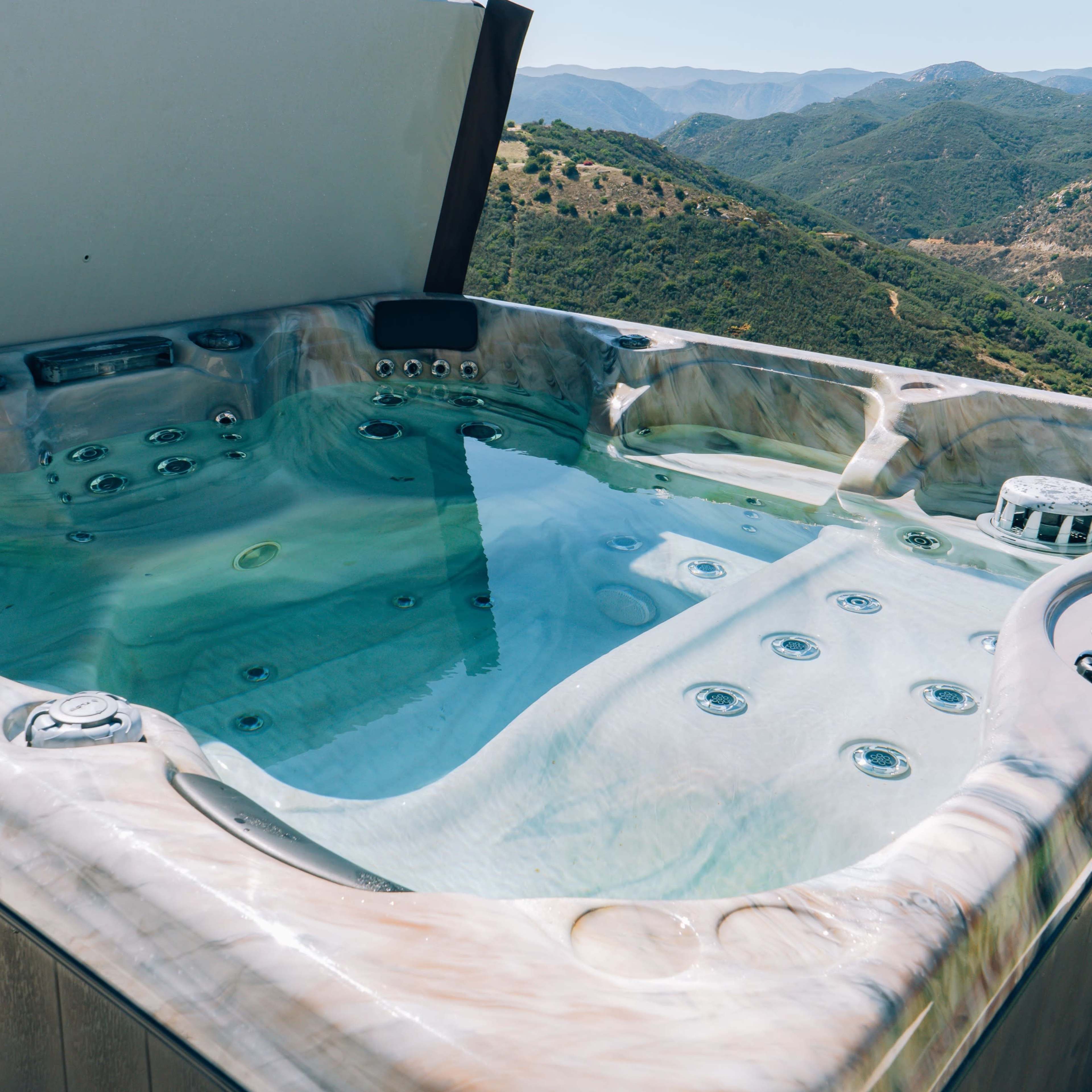 A hot tub filled with water sits on a deck overlooking rolling hills and mountains.