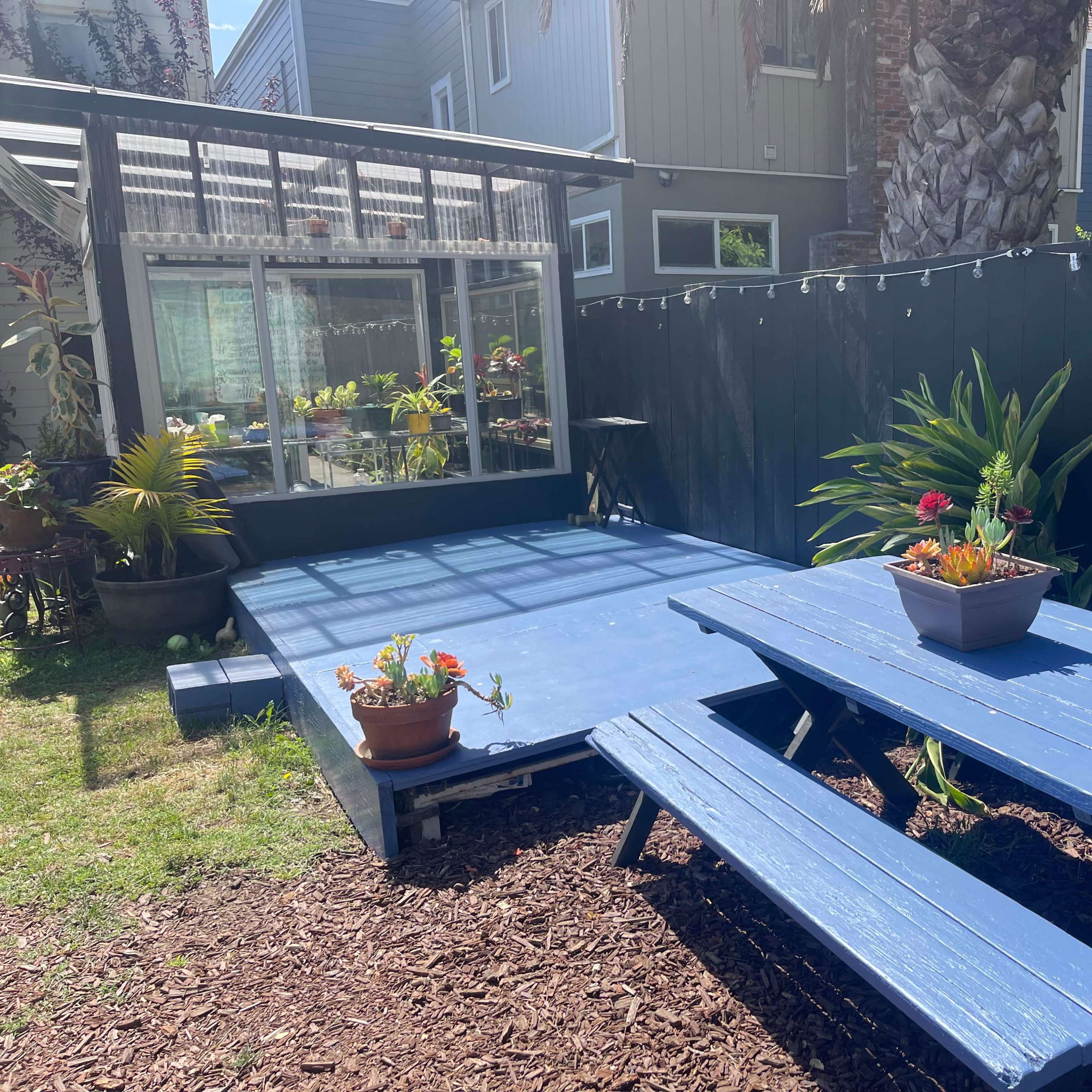 A blue wooden deck with a picnic table and potted plants is set in a backyard next to a greenhouse and surrounded by a black fence.