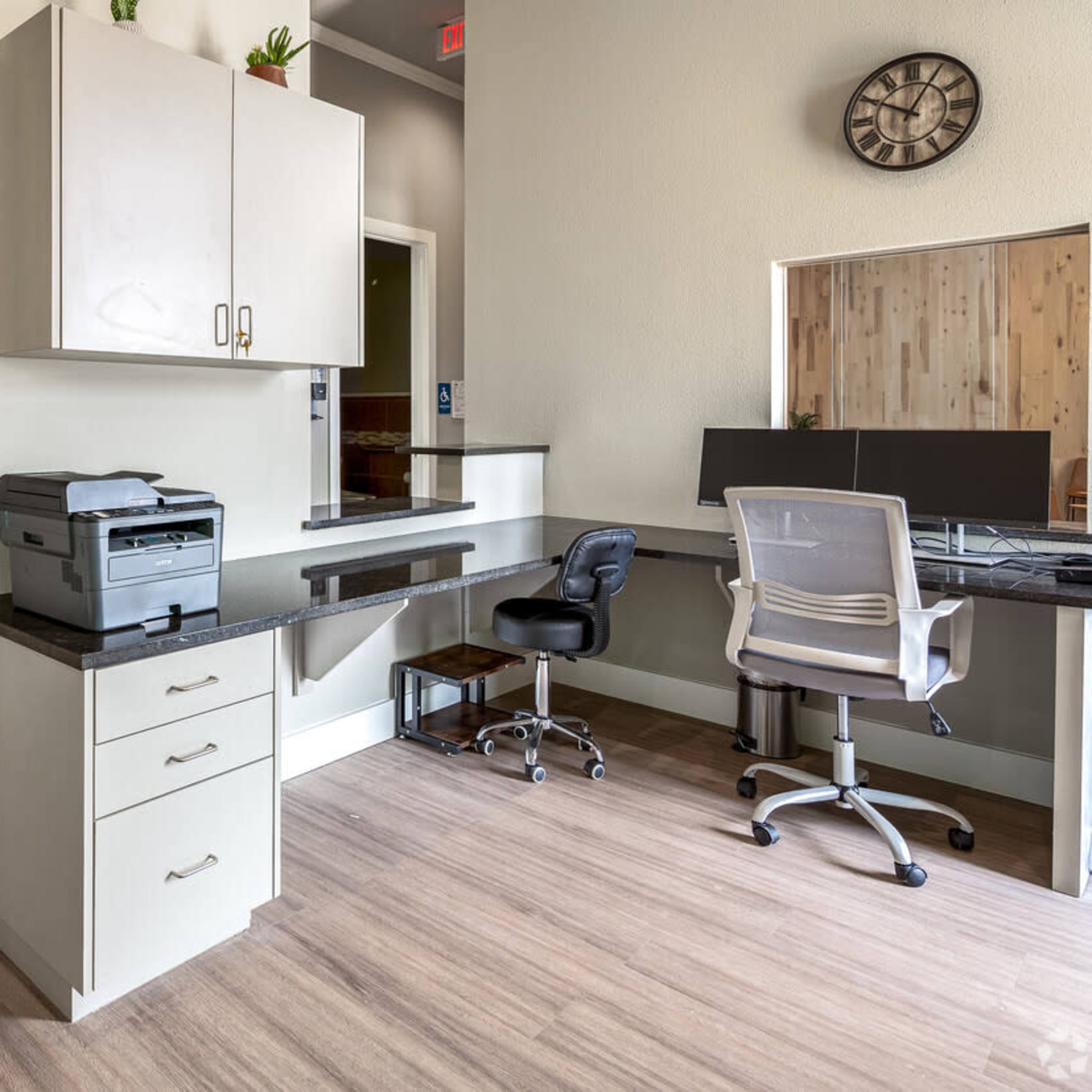 A modern office space featuring a printer on a countertop, a desk with a computer setup, and light wood flooring.