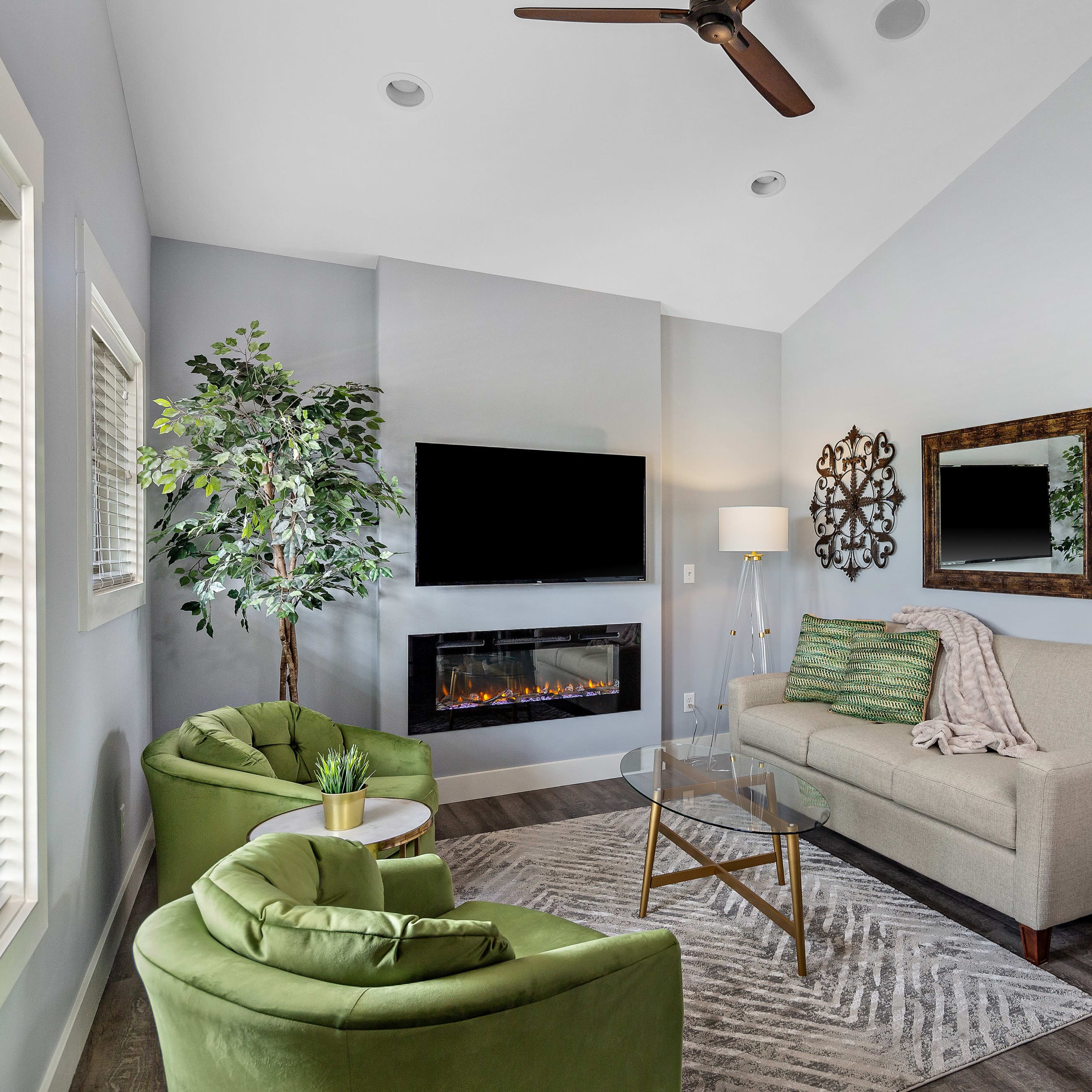 A modern living room featuring a green sofa and armchairs, a wall-mounted television, an electric fireplace, and decorative mirrors on the walls.