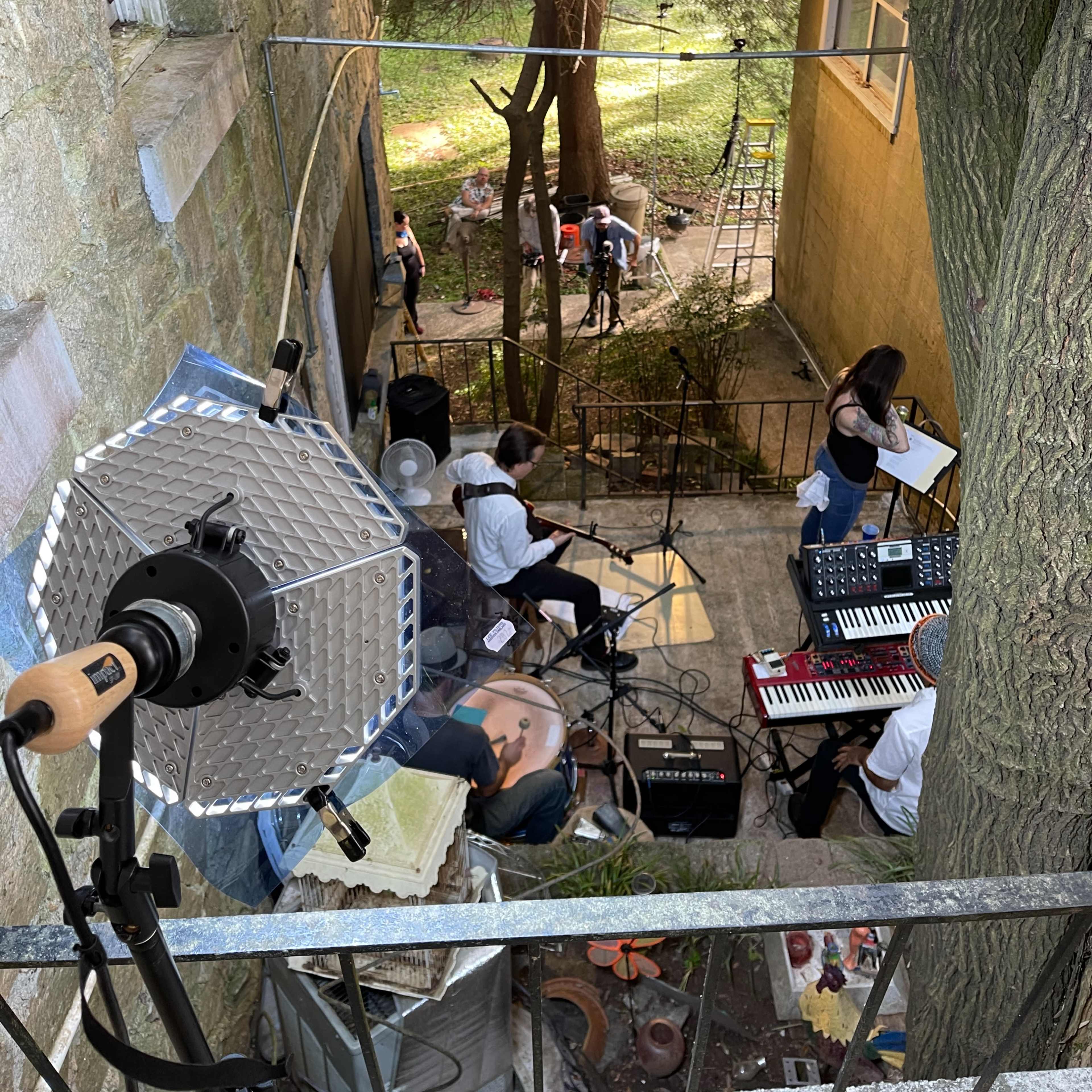 A group of musicians performs in a courtyard surrounded by trees and a building, with a spotlight illuminating the area.