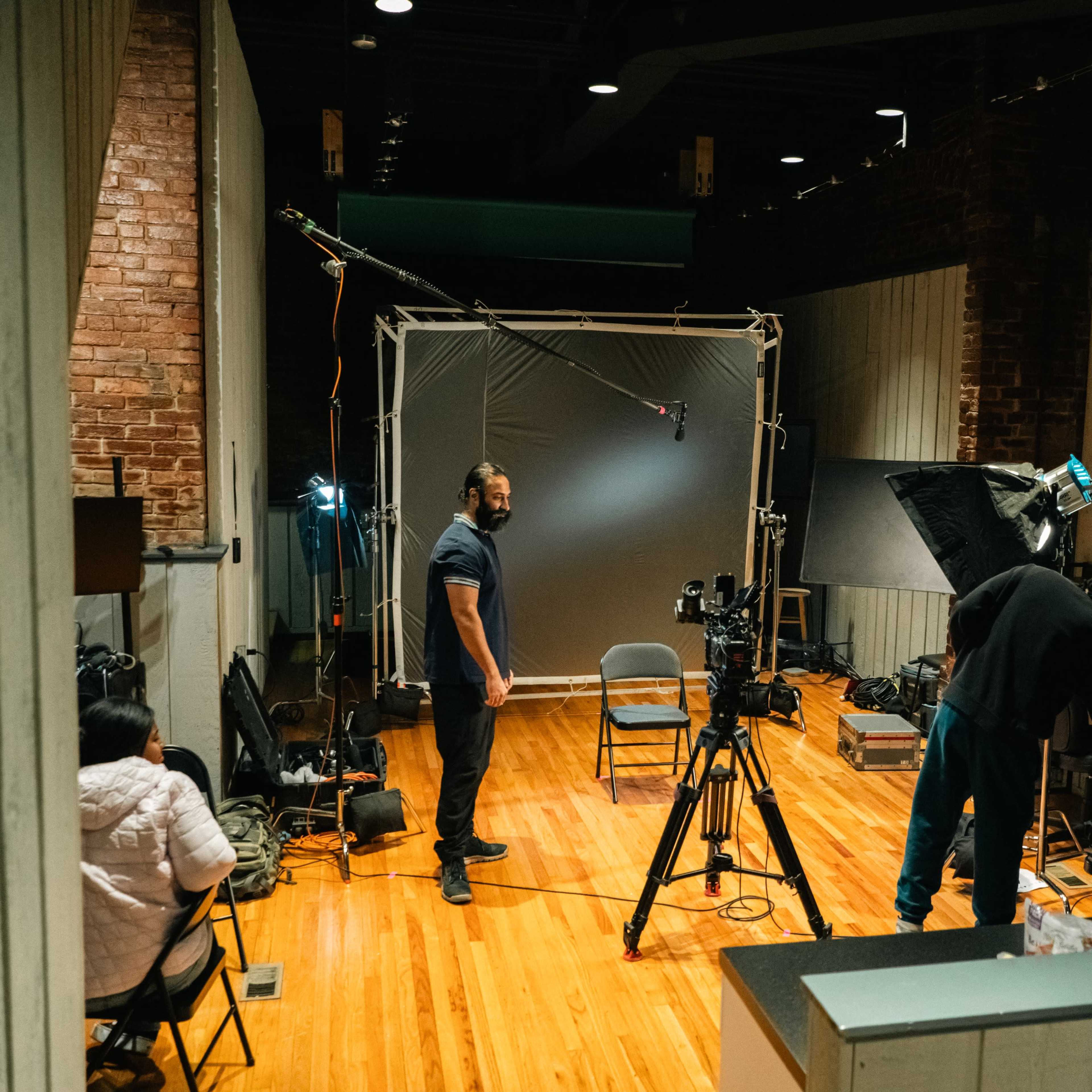 A video production studio features two people preparing equipment near a backdrop and camera setup.