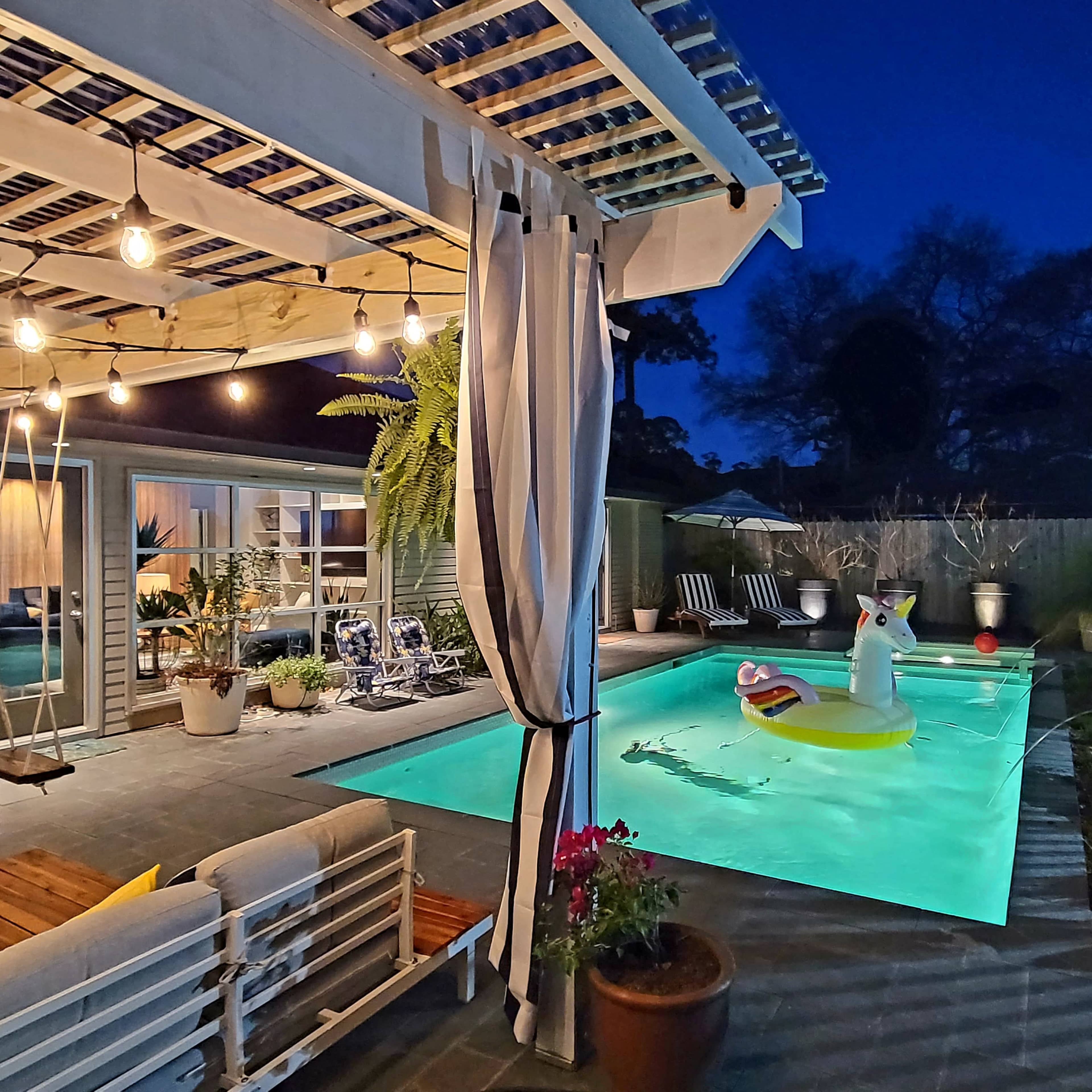 The scene depicts a well-lit backyard pool area at night, featuring a turquoise pool with a large inflatable unicorn and surrounded by comfortable seating and decorative plants.