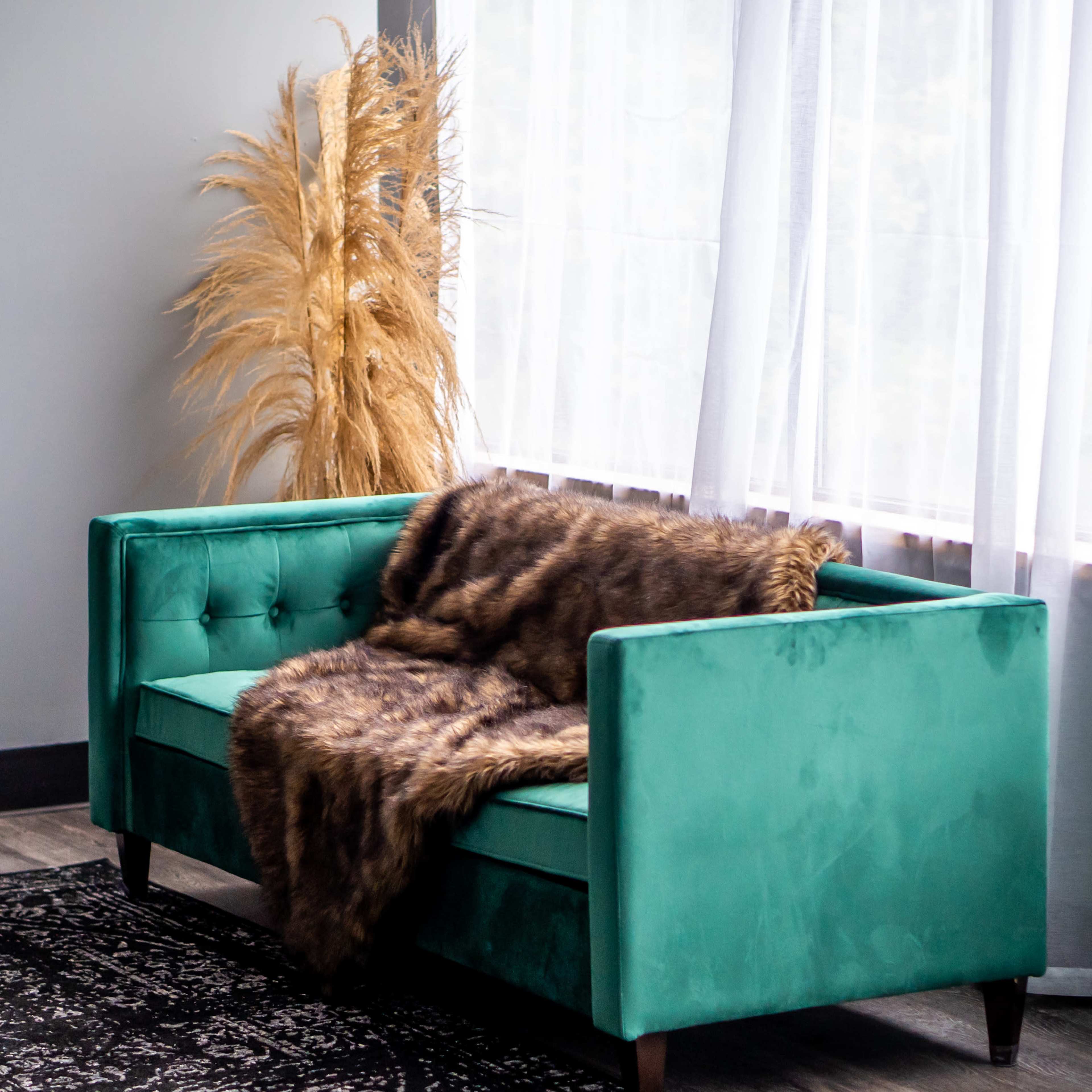 A teal velvet sofa with a fur throw rests against a large window covered by sheer curtains, alongside a decorative arrangement of tall dried plants.