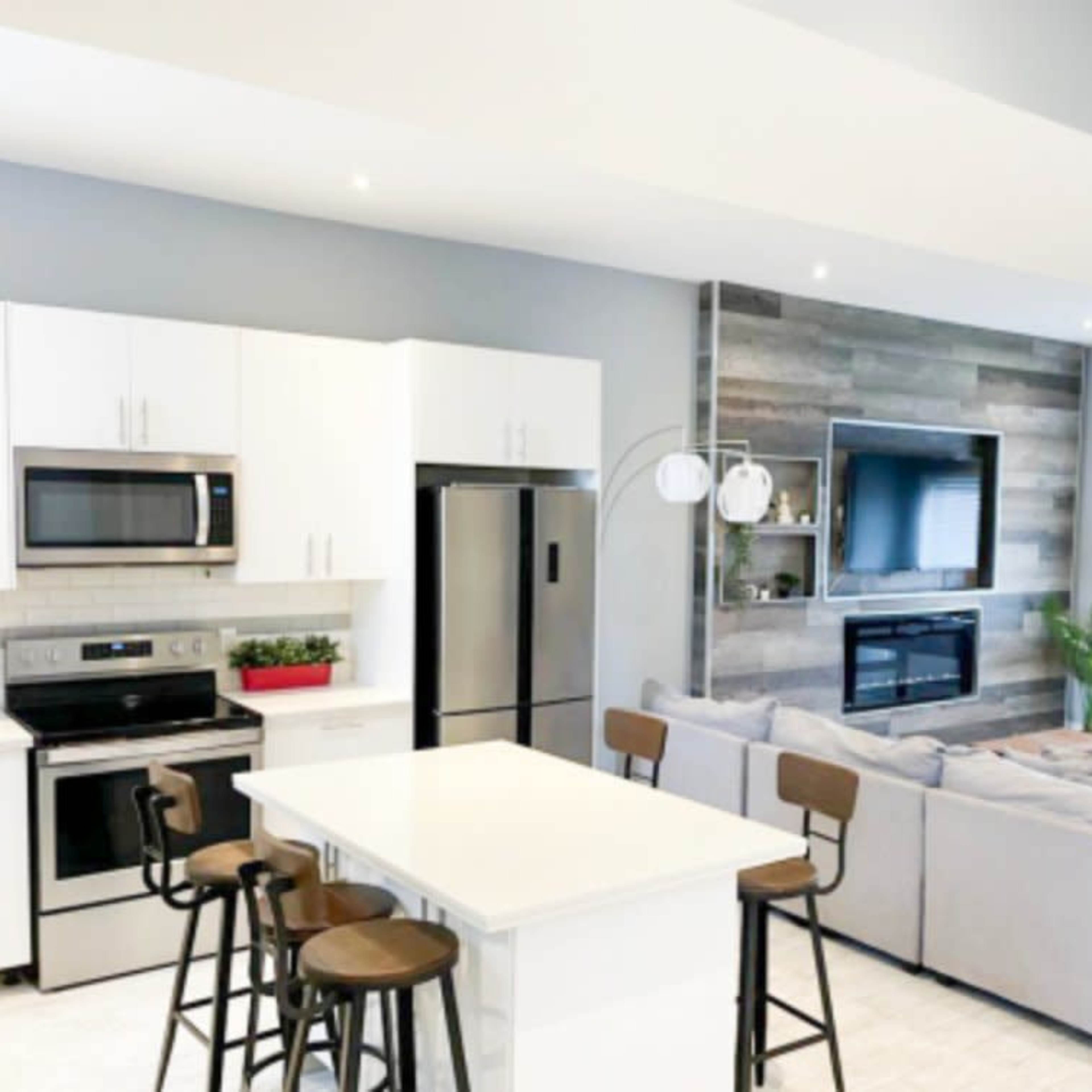 A modern kitchen and living area featuring white cabinets, stainless steel appliances, a large sectional couch, and a wall-mounted television above a fireplace.
