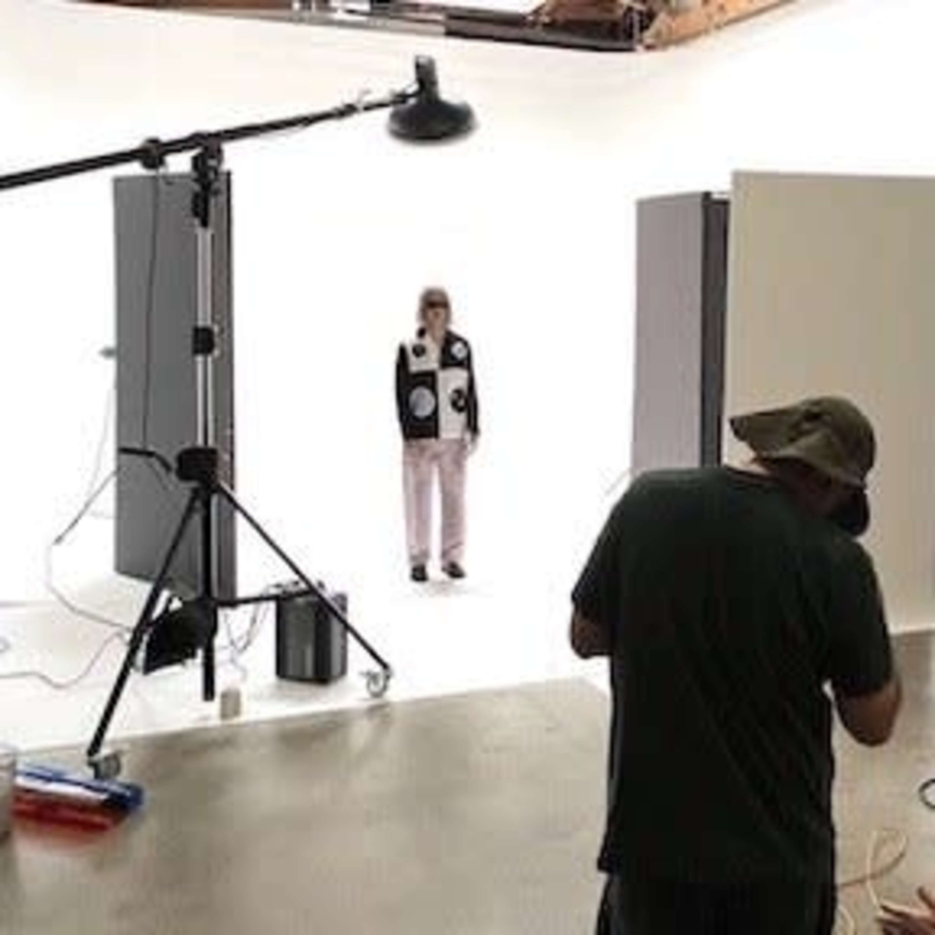 A photographer is setting up a shoot with a model standing in front of a plain white backdrop in a studio.