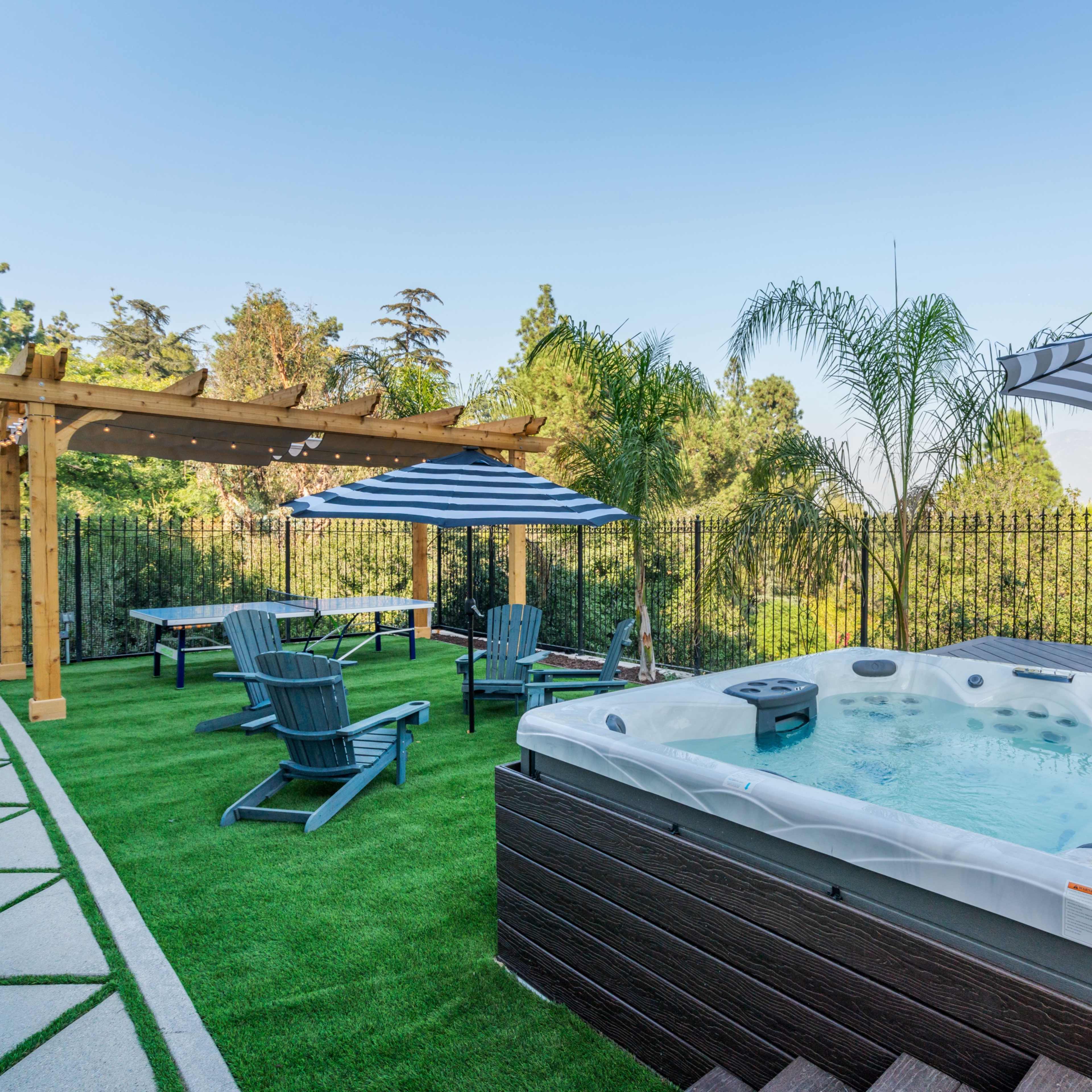 A backyard with a hot tub, patio furniture under striped umbrellas, and a landscaped area featuring green grass and decorative trees.