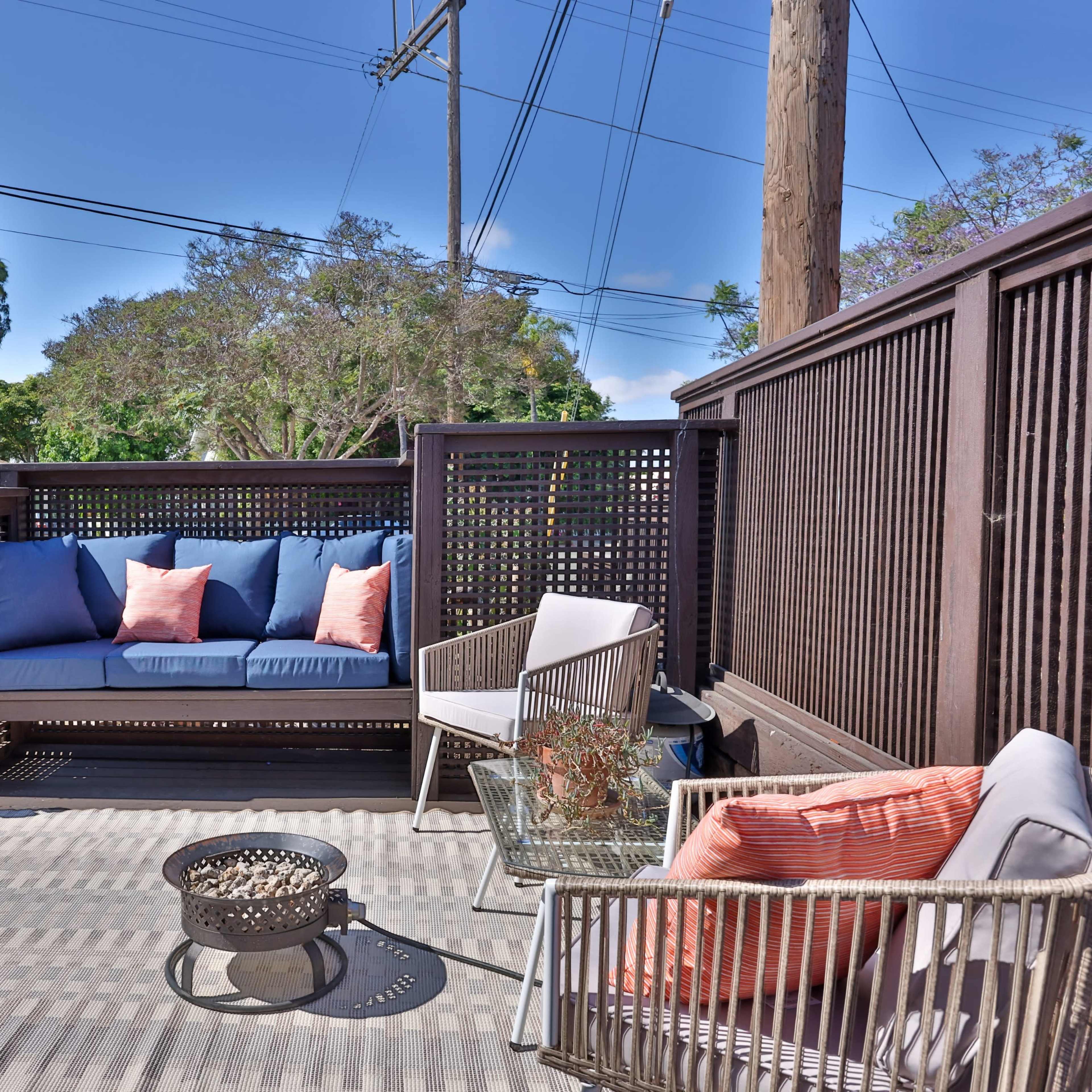 A cozy outdoor patio with a sofa, two chairs, a small fire pit, and surrounding greenery.
