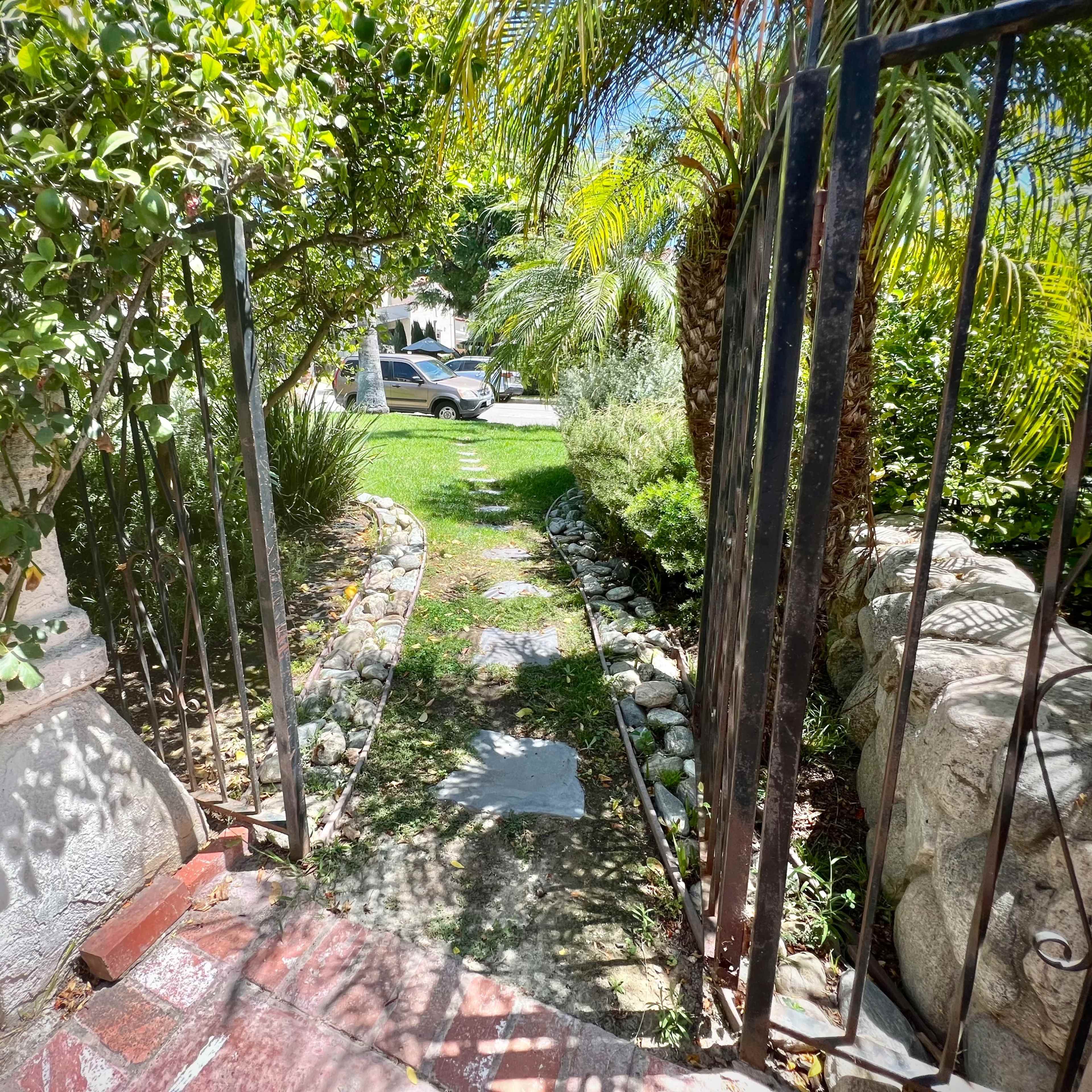 A narrow stone pathway leads through a garden gate, bordered by greenery and rocks, toward a lawn visible in the distance.