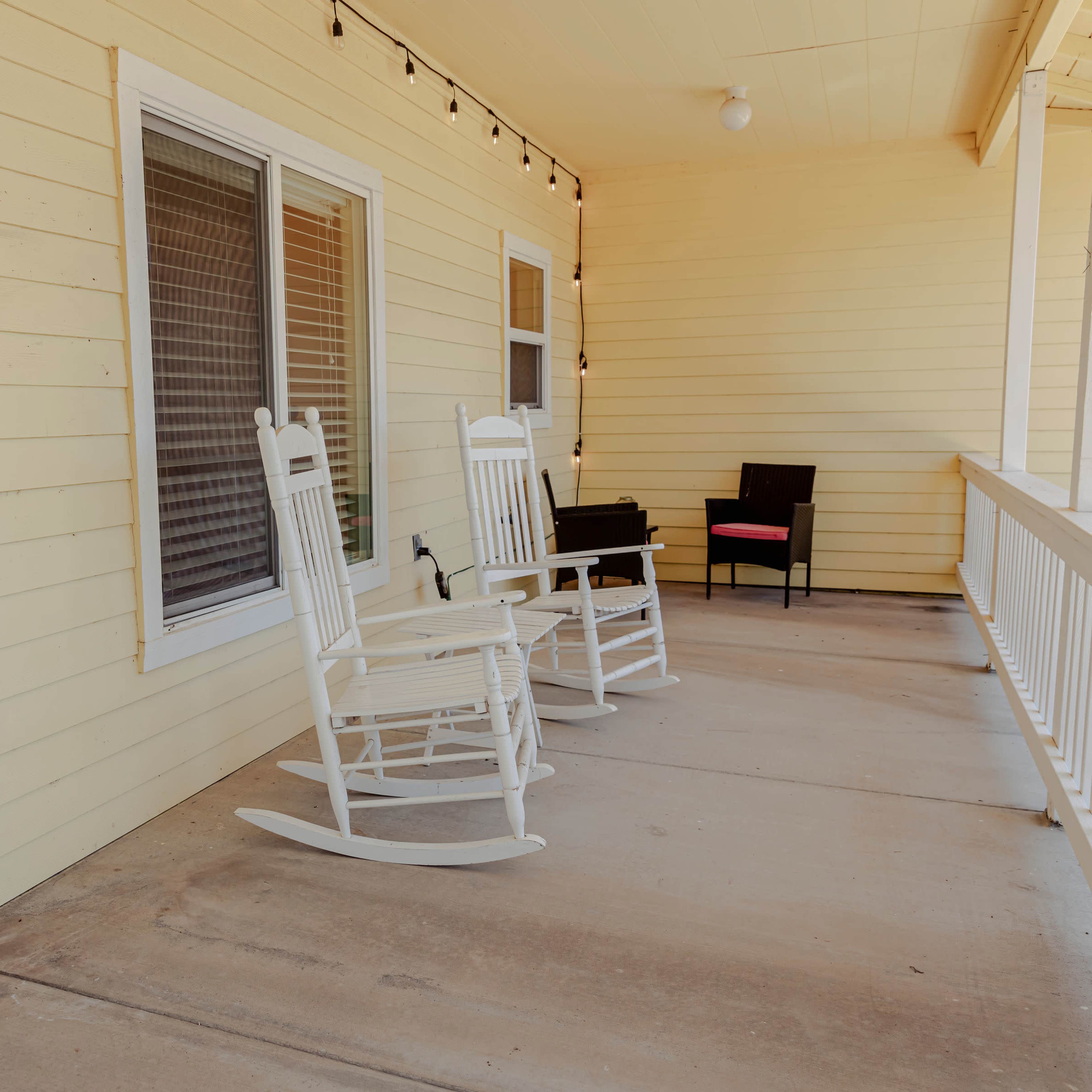 A covered porch features two white rocking chairs and a small black seat beside a welcoming sign on the wall.