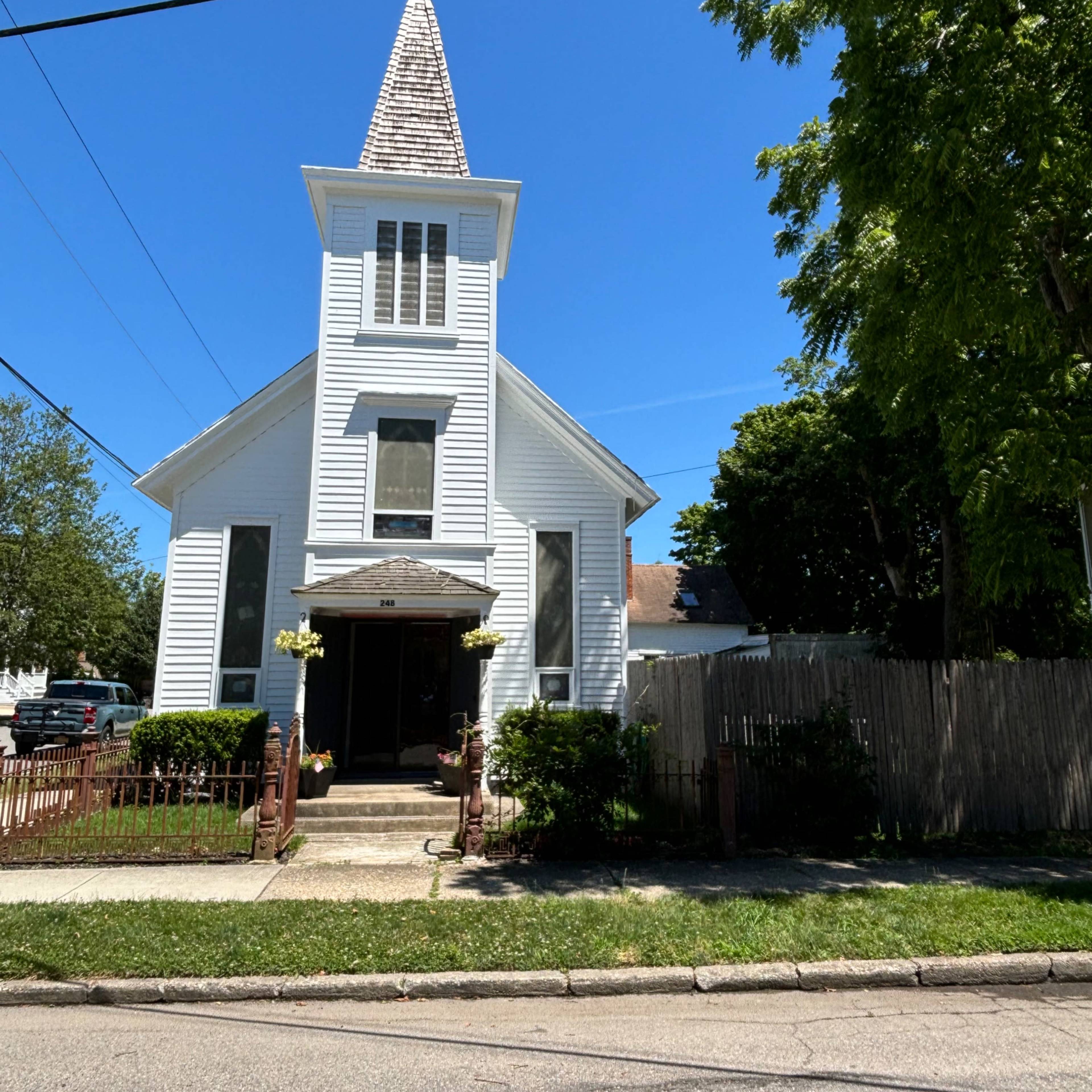 A white, wooden church with a tall steeple and cross sits at the corner of a street, surrounded by greenery.